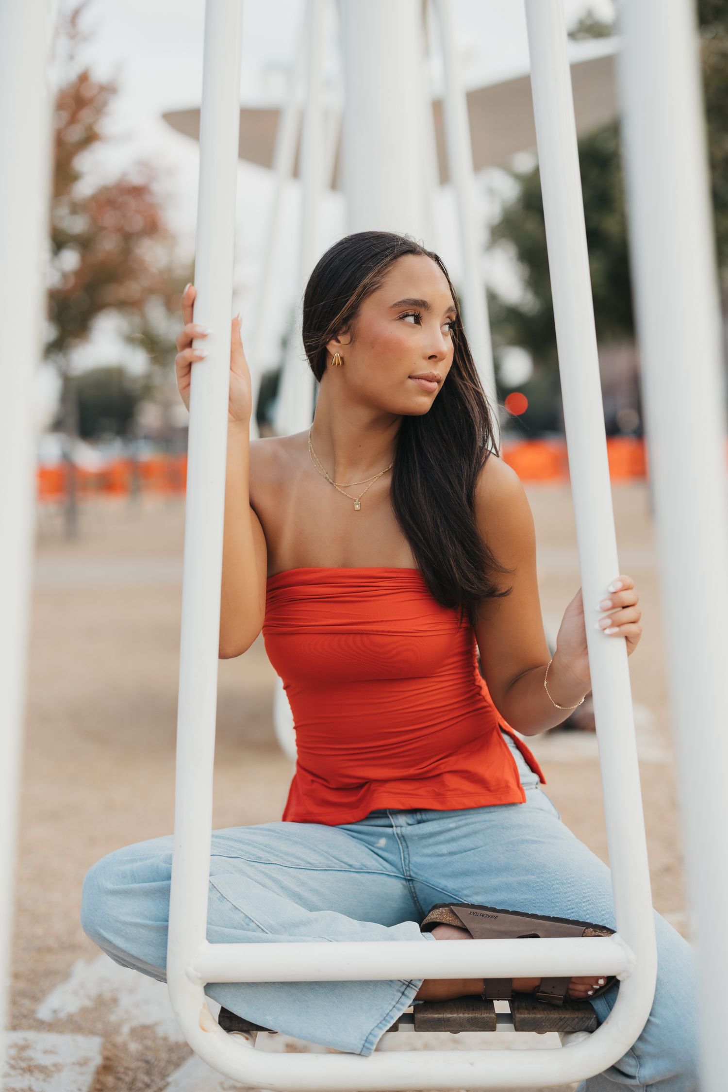 Young woman in red tube top and jeans sitting on white swing outdoors, looking away thoughtfully.