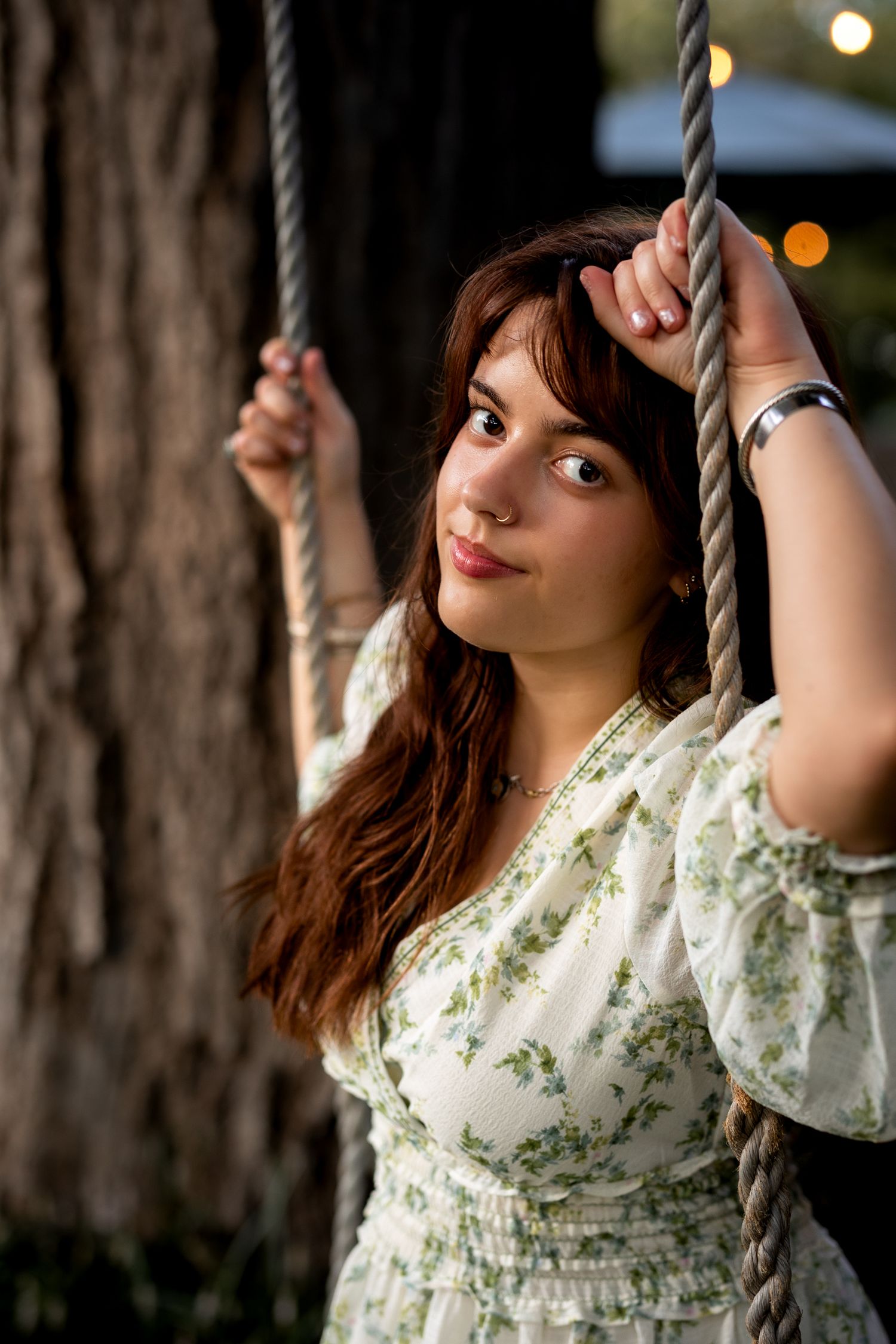 Young woman in floral dress holding rope swing near tree, smiling softly at camera outdoors.