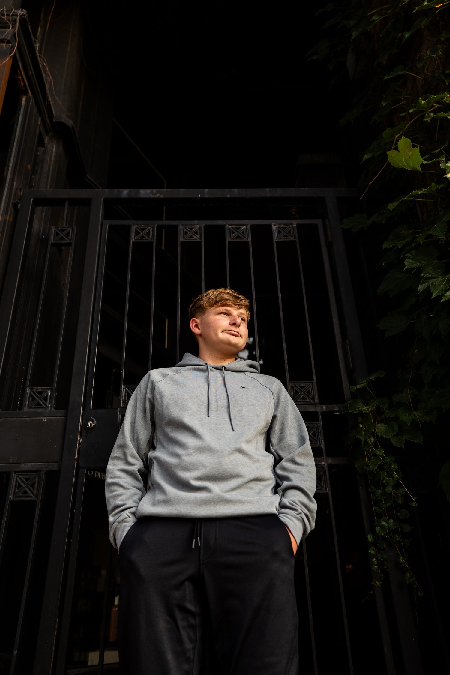 Young man in gray Nike hoodie and black joggers standing confidently before dark iron gates with green foliage.