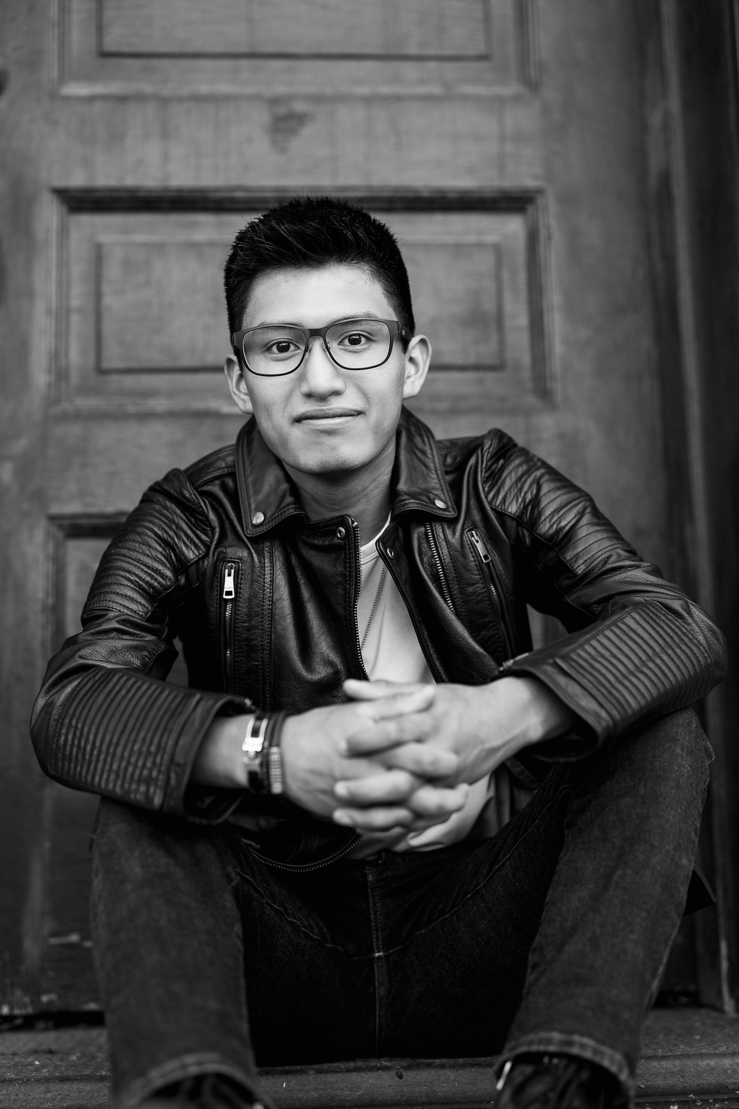 Person in a leather jacket and glasses poses against dark wooden door background in a series.
