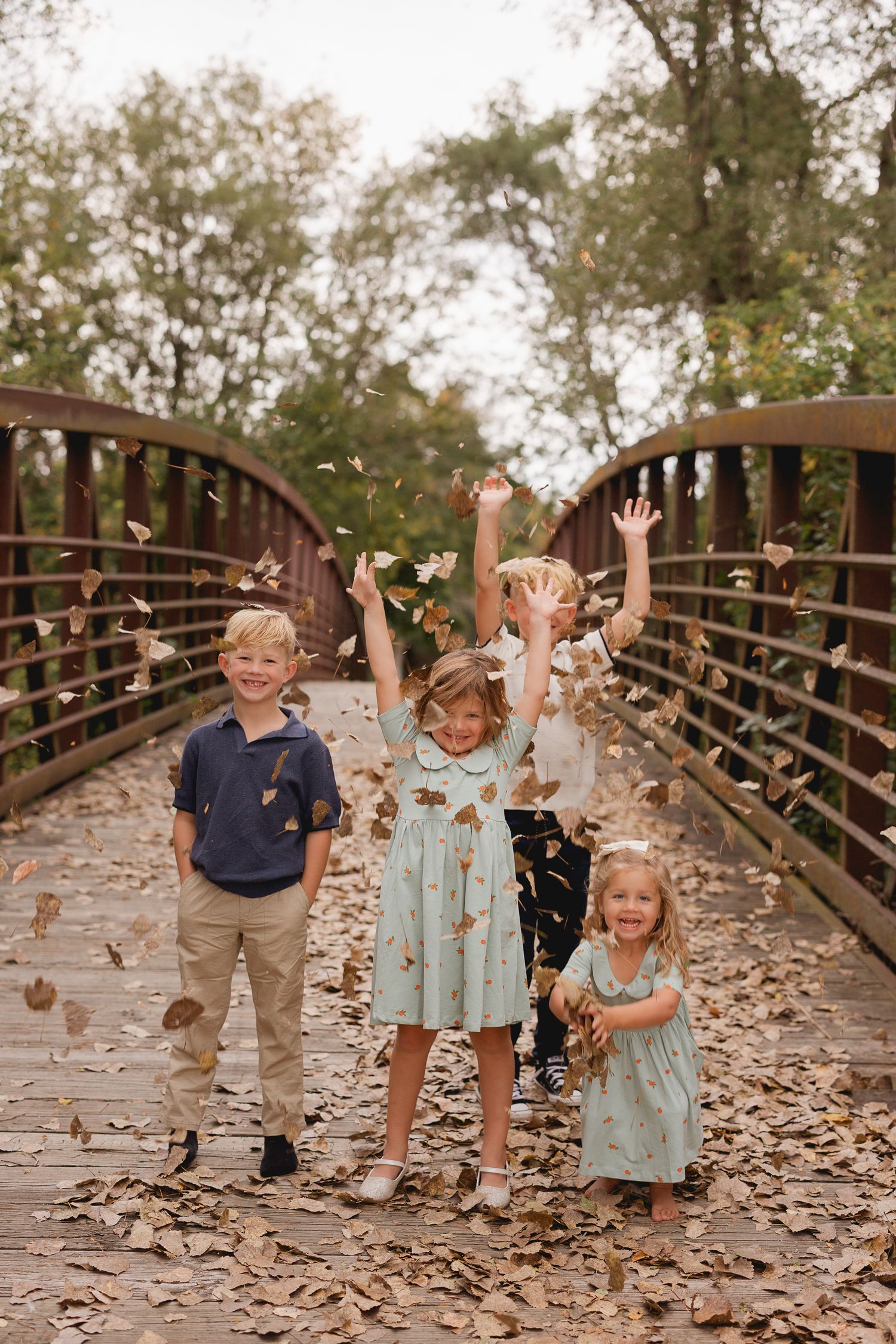 Four joyful children toss autumn leaves in the air on a wooden bridge surrounded by fall foliage.