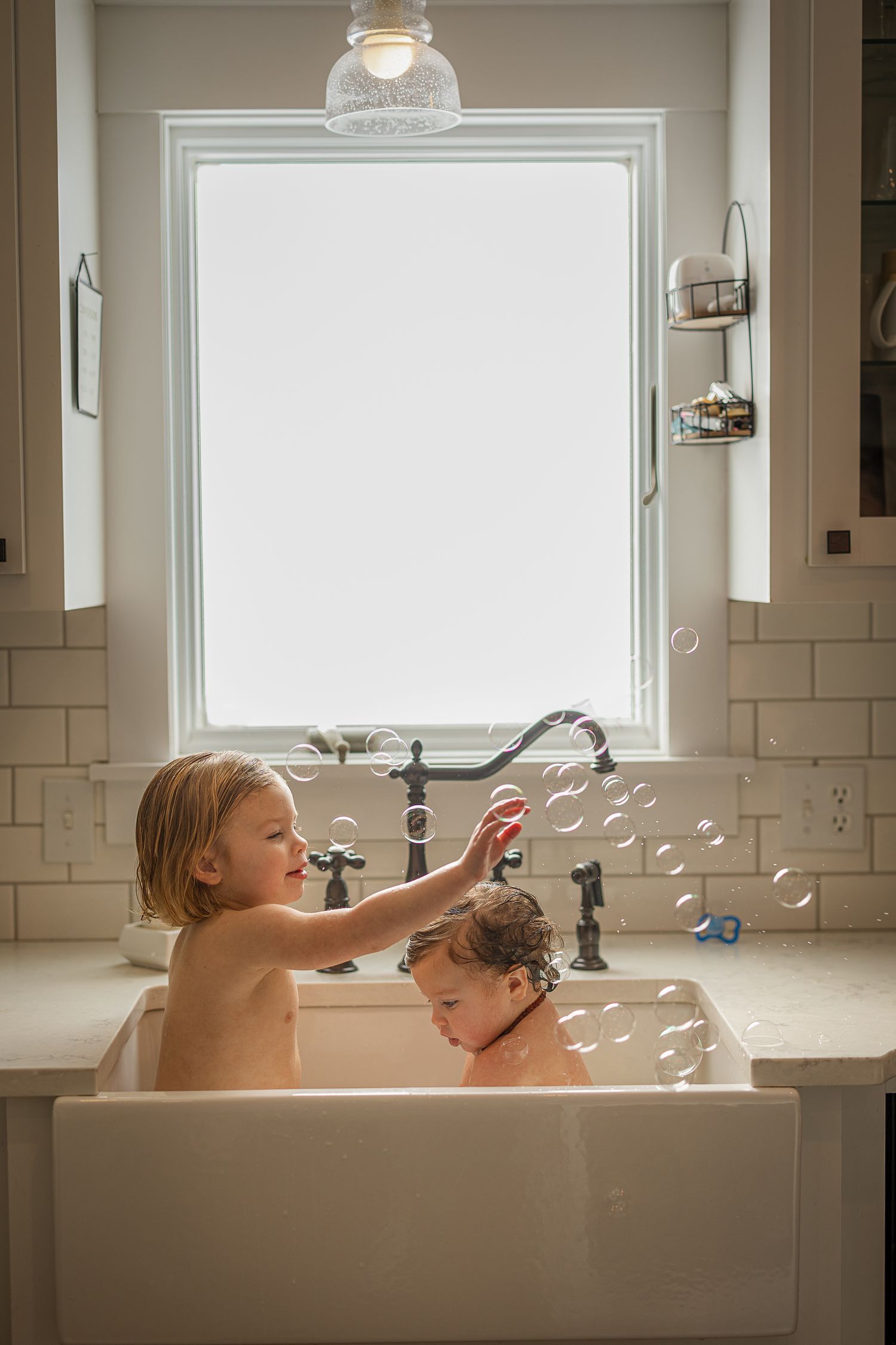 Two young children playing with bubbles while bathing together in a farmhouse kitchen sink near a bright window.