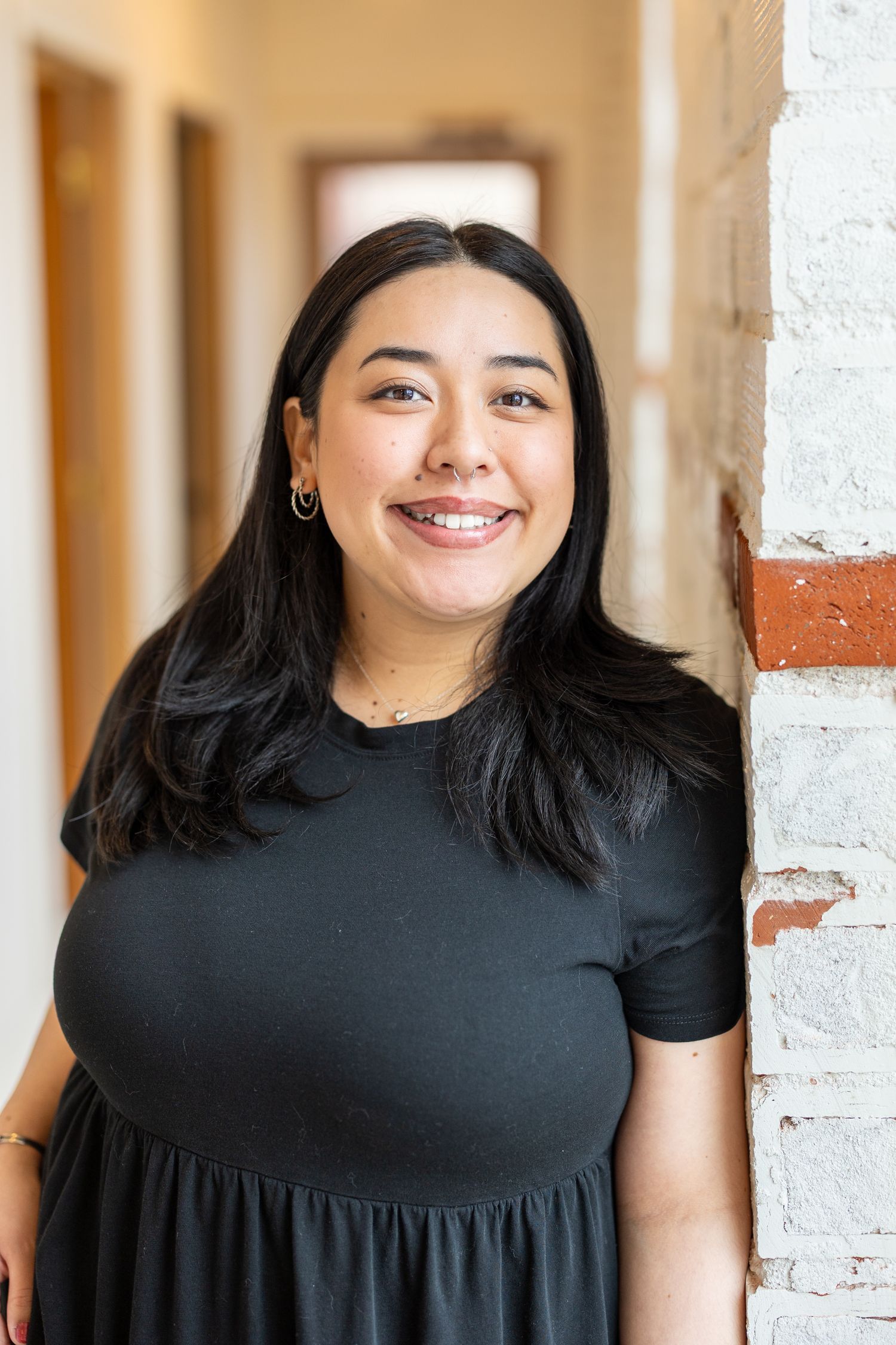 Smiling young woman with long black hair wearing a black dress, leaning against a white brick wall in a hallway.