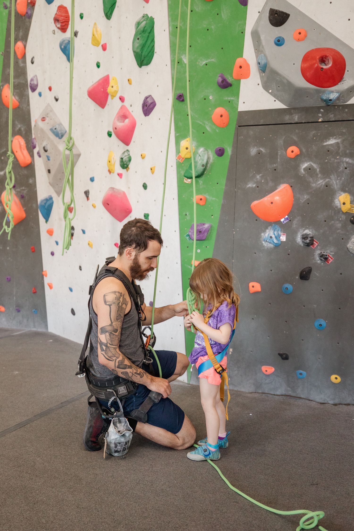 Tattooed instructor kneeling to help young girl with climbing harness at indoor rock climbing gym.