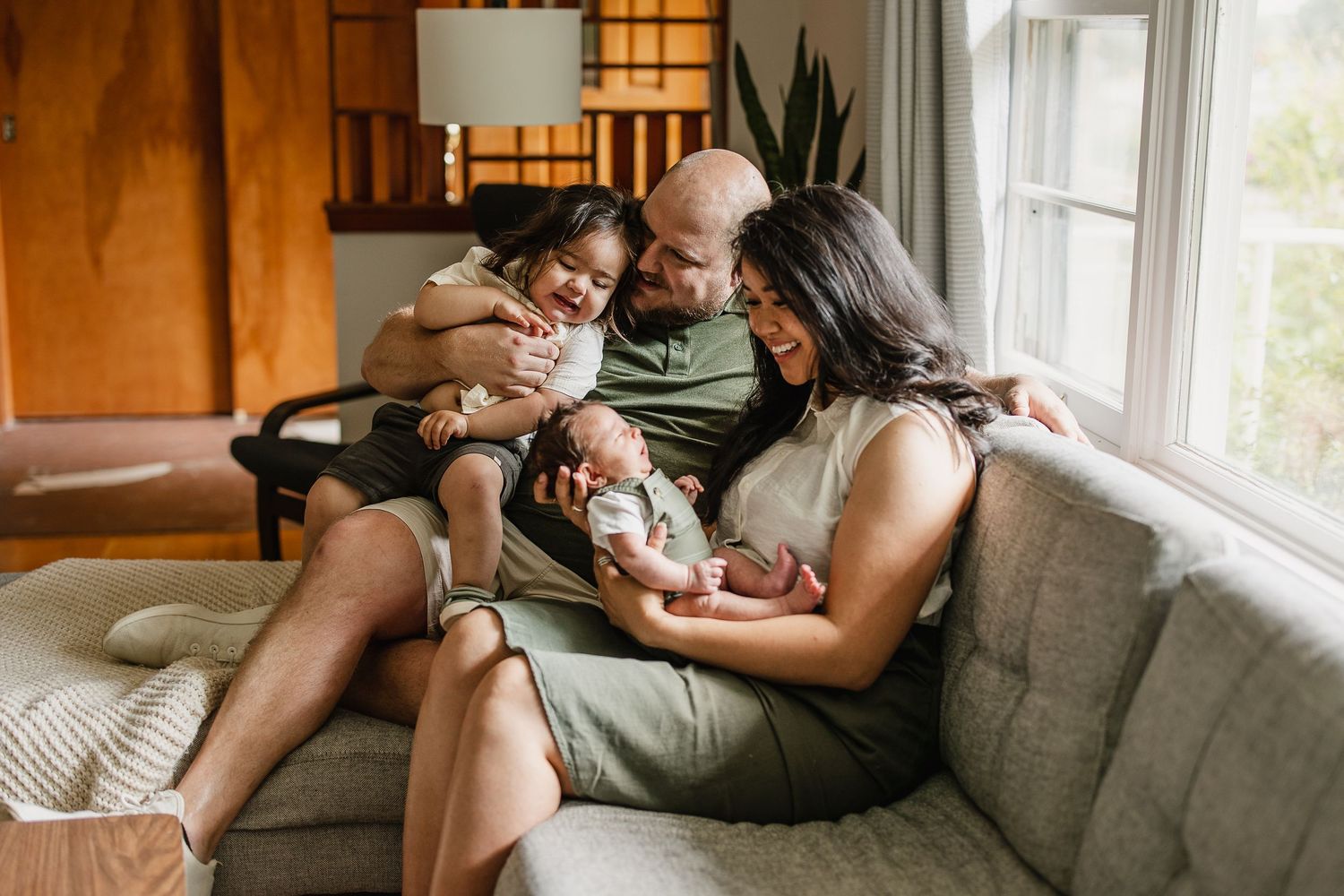 Happy family of four sitting on a cozy sofa, parents lovingly holding their toddler and newborn baby at home.