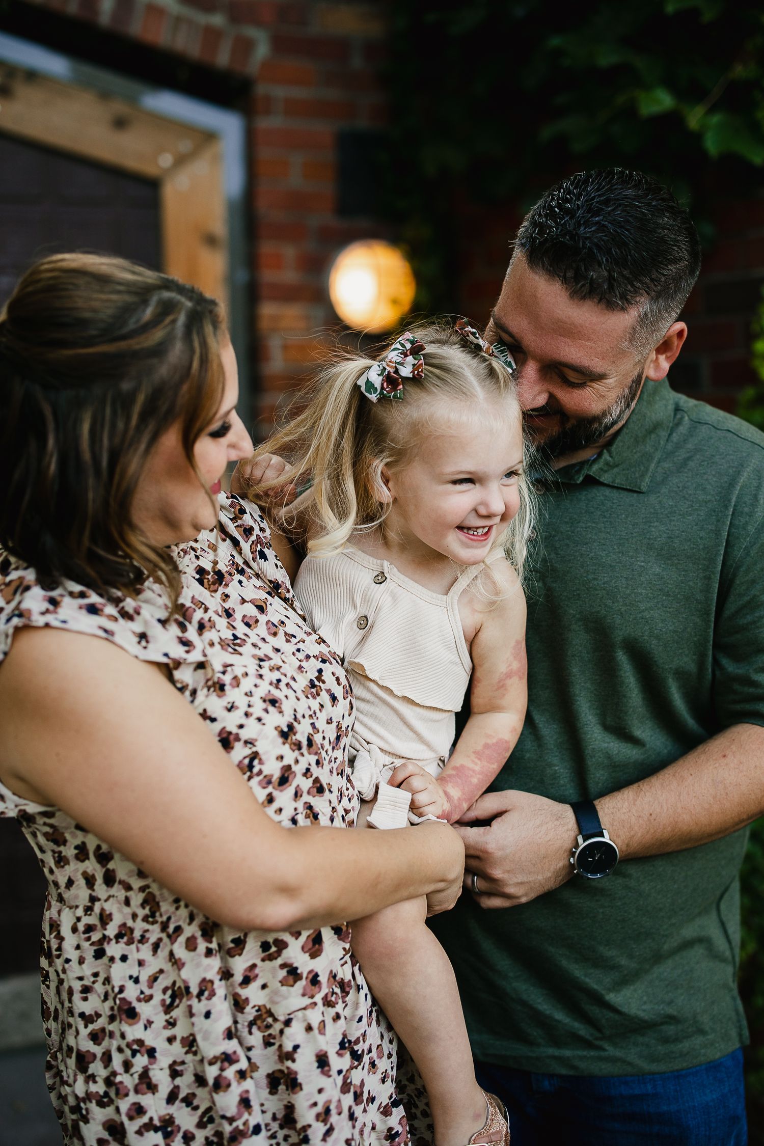 A joyful family of three with parents holding their smiling blonde toddler girl outdoors near a brick wall.