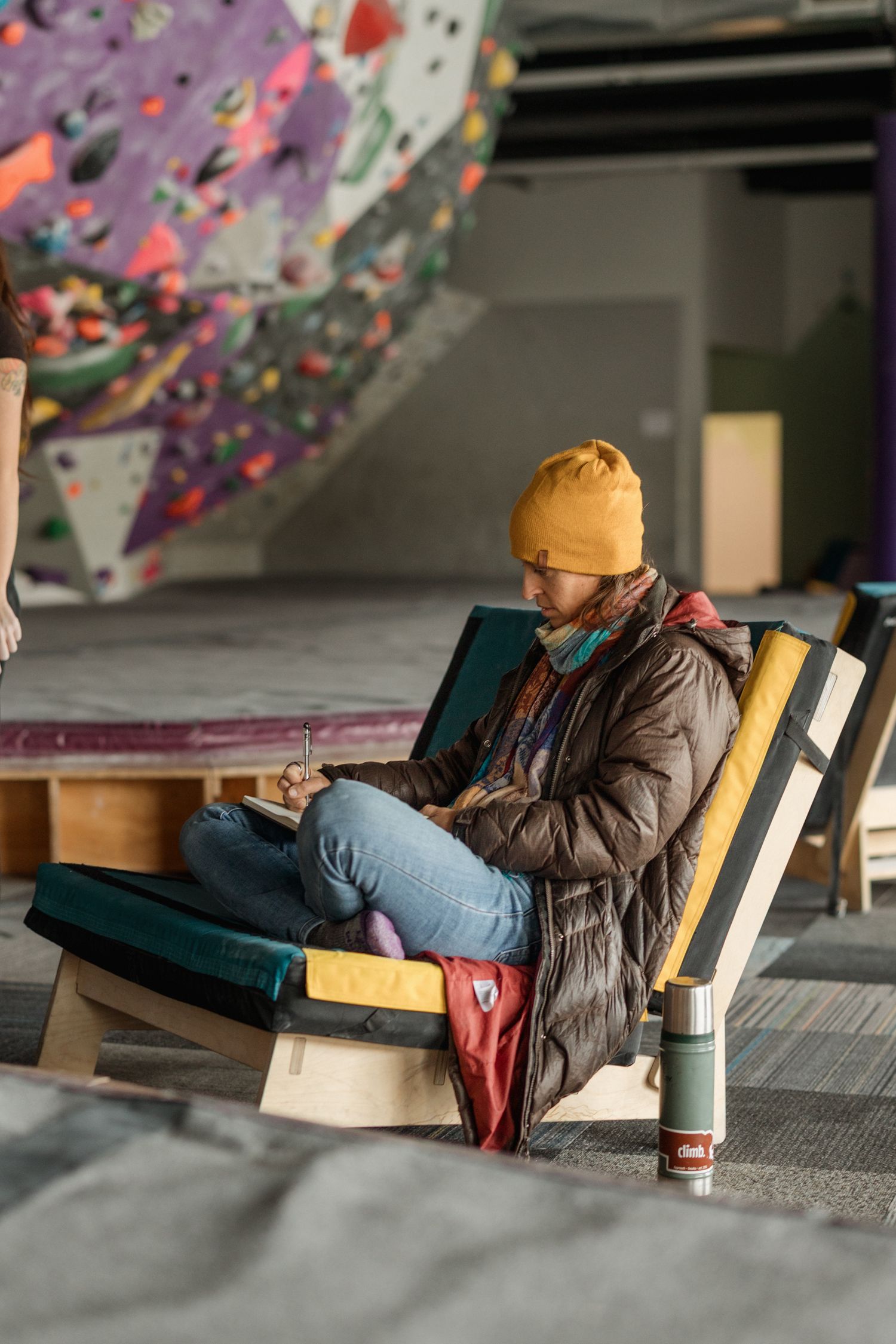 Woman in yellow beanie writing in notebook while sitting in wooden chair at indoor rock climbing gym.