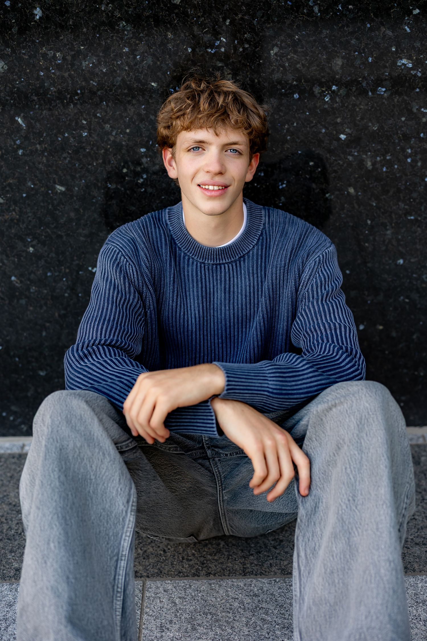 Smiling teenage boy in blue ribbed sweater and jeans sitting on granite steps against dark marble wall.