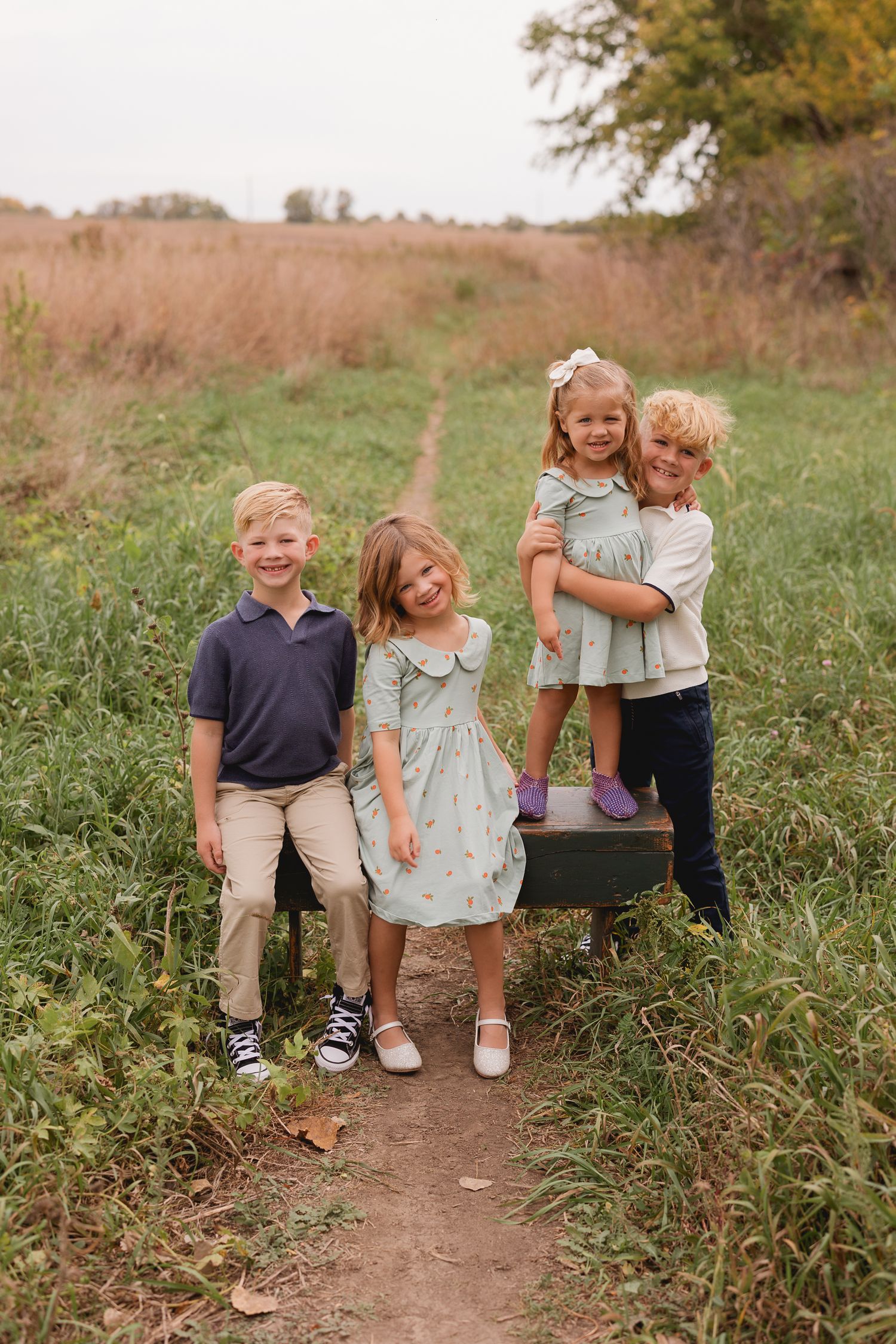 Four blonde children pose together on a dirt path in an autumn field, dressed in coordinating sage green and navy outfits.