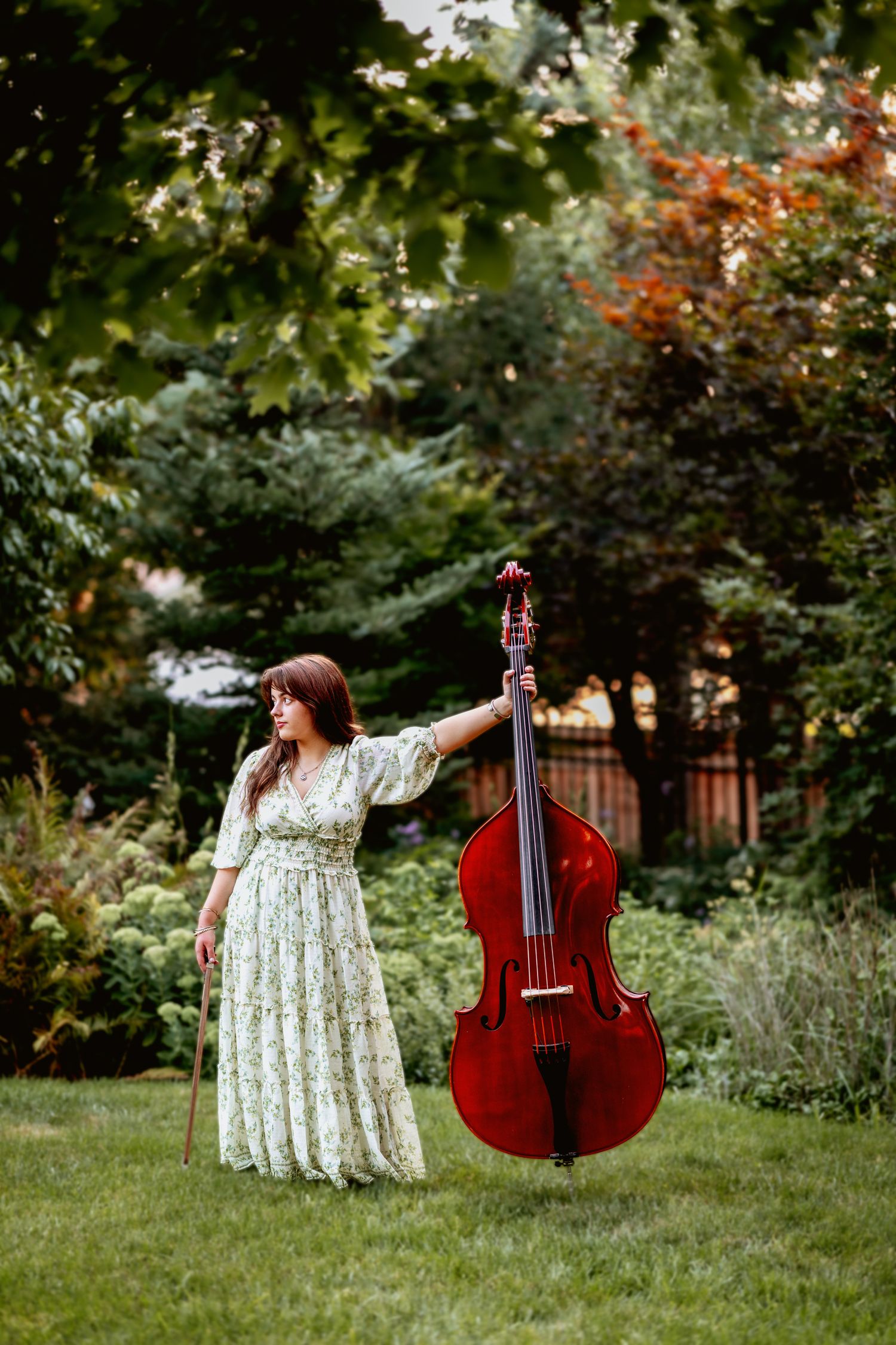 Woman in floral dress holding a double bass in a lush green garden setting.