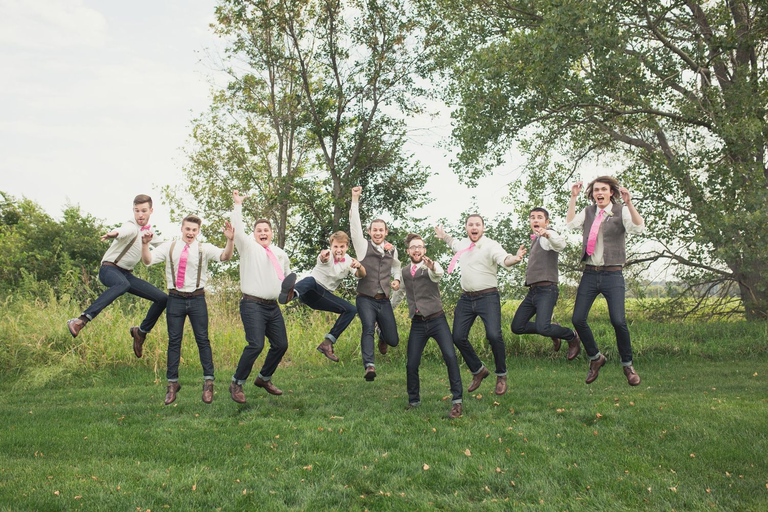 Groomsmen in gray suits and pink ties jumping joyfully on green grass with trees in background at outdoor wedding.