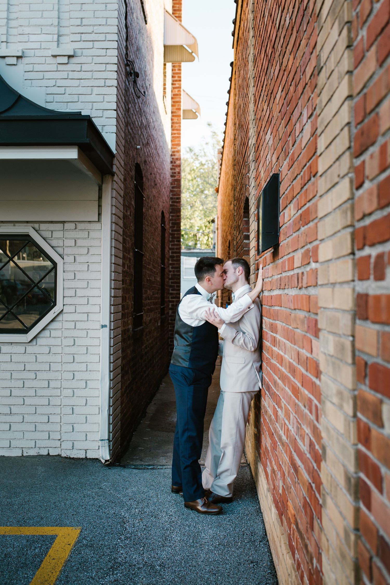 Two men sharing a romantic kiss in a narrow brick alley, one in a gray vest and one in a cream suit.