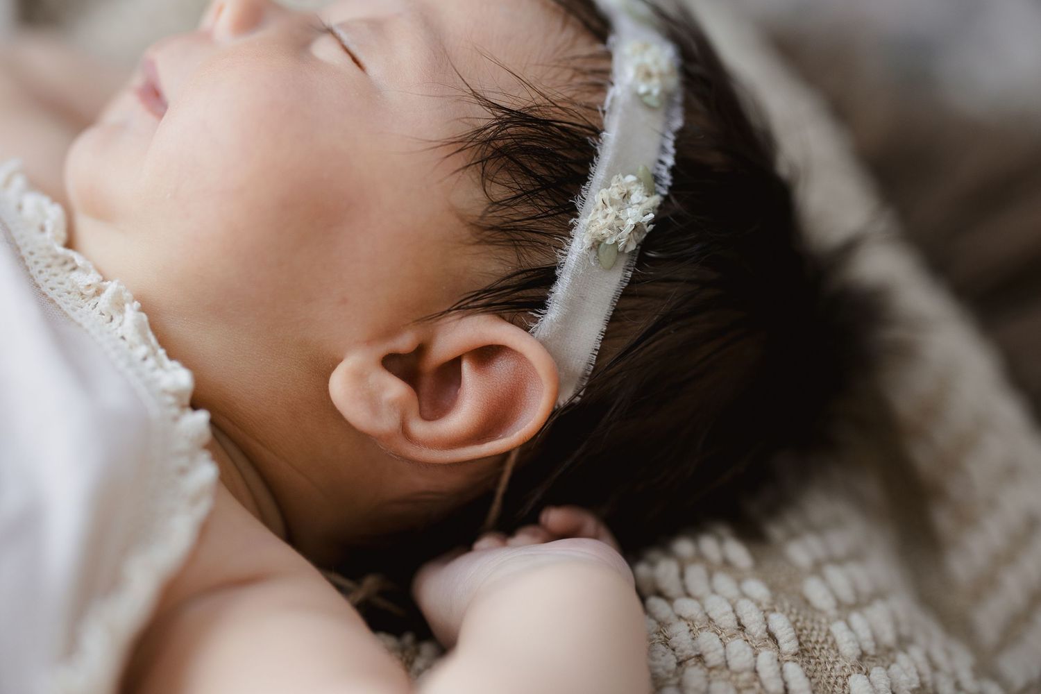 Peaceful newborn baby sleeping with dark hair and a delicate floral headband, wearing a white lace outfit.