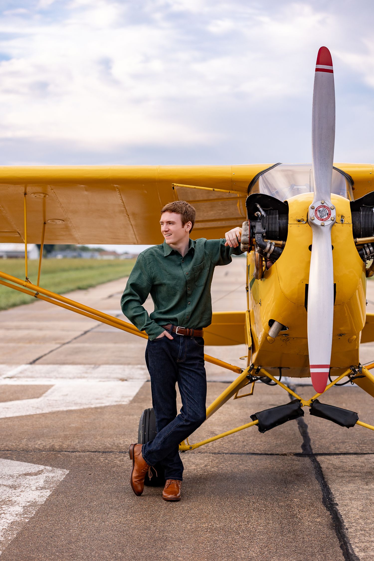 Young man in green shirt leaning on yellow propeller plane on airstrip.