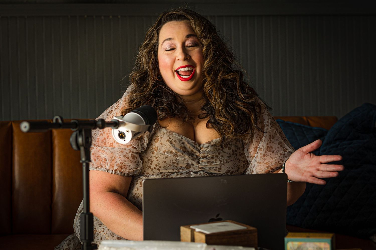 A woman laughs joyfully while sitting with a laptop and puppet microphone in a warmly lit studio setting.