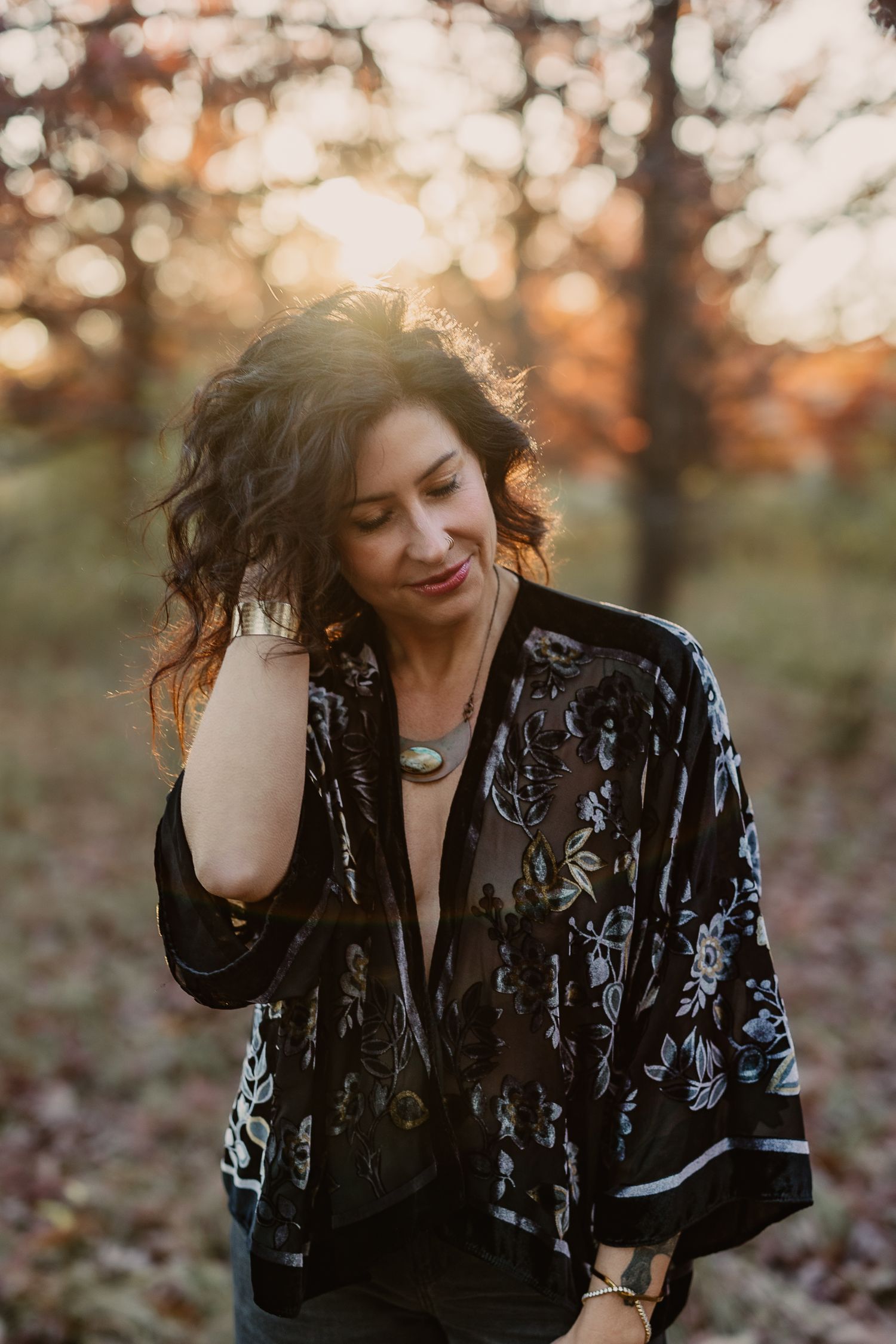 Woman in black floral kimono poses outdoors in golden autumn light with bokeh forest background.