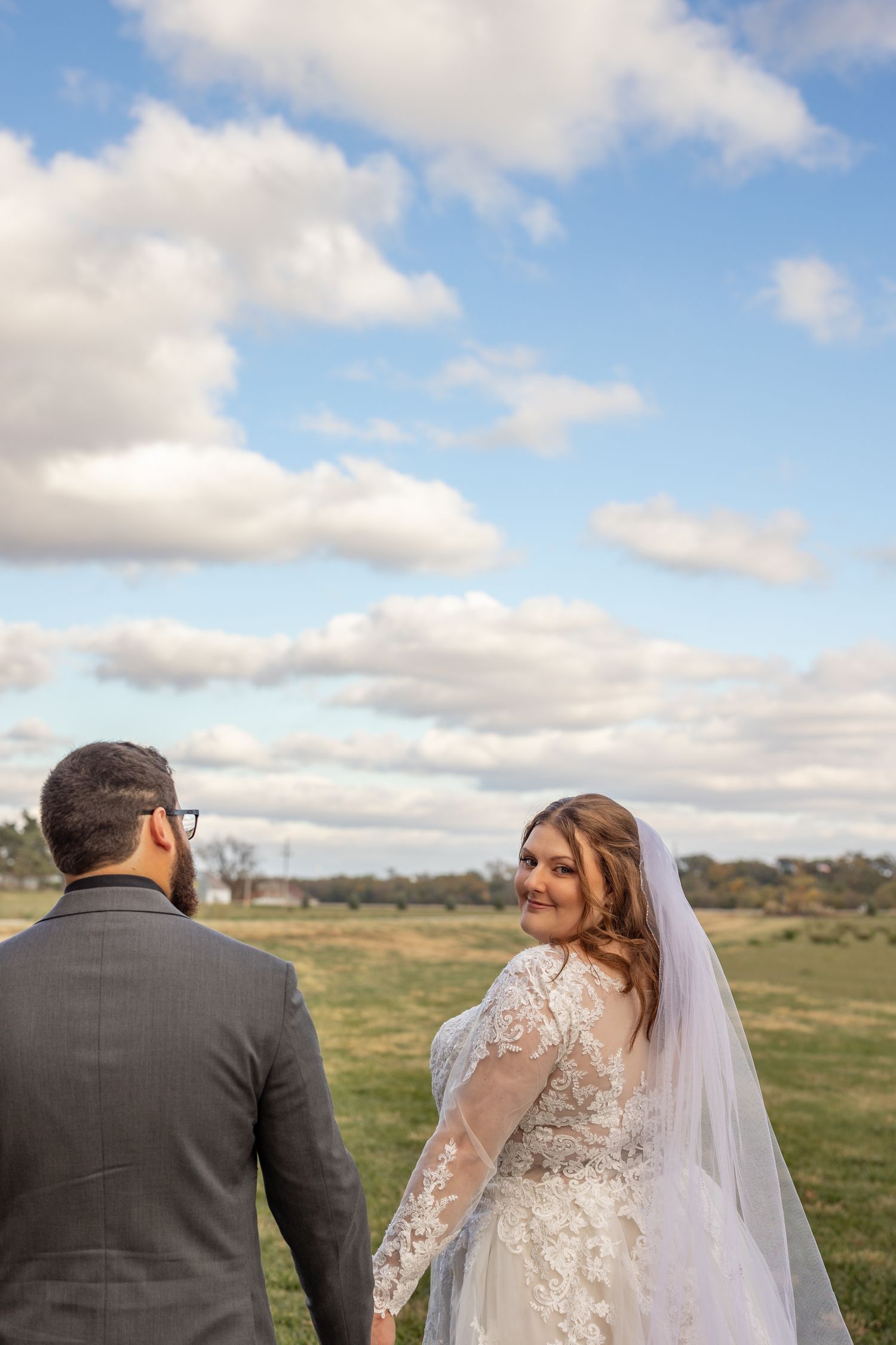 Bride in lace gown and veil holds groom's hand, smiling over shoulder at camera with green fields and cloudy sky behind.