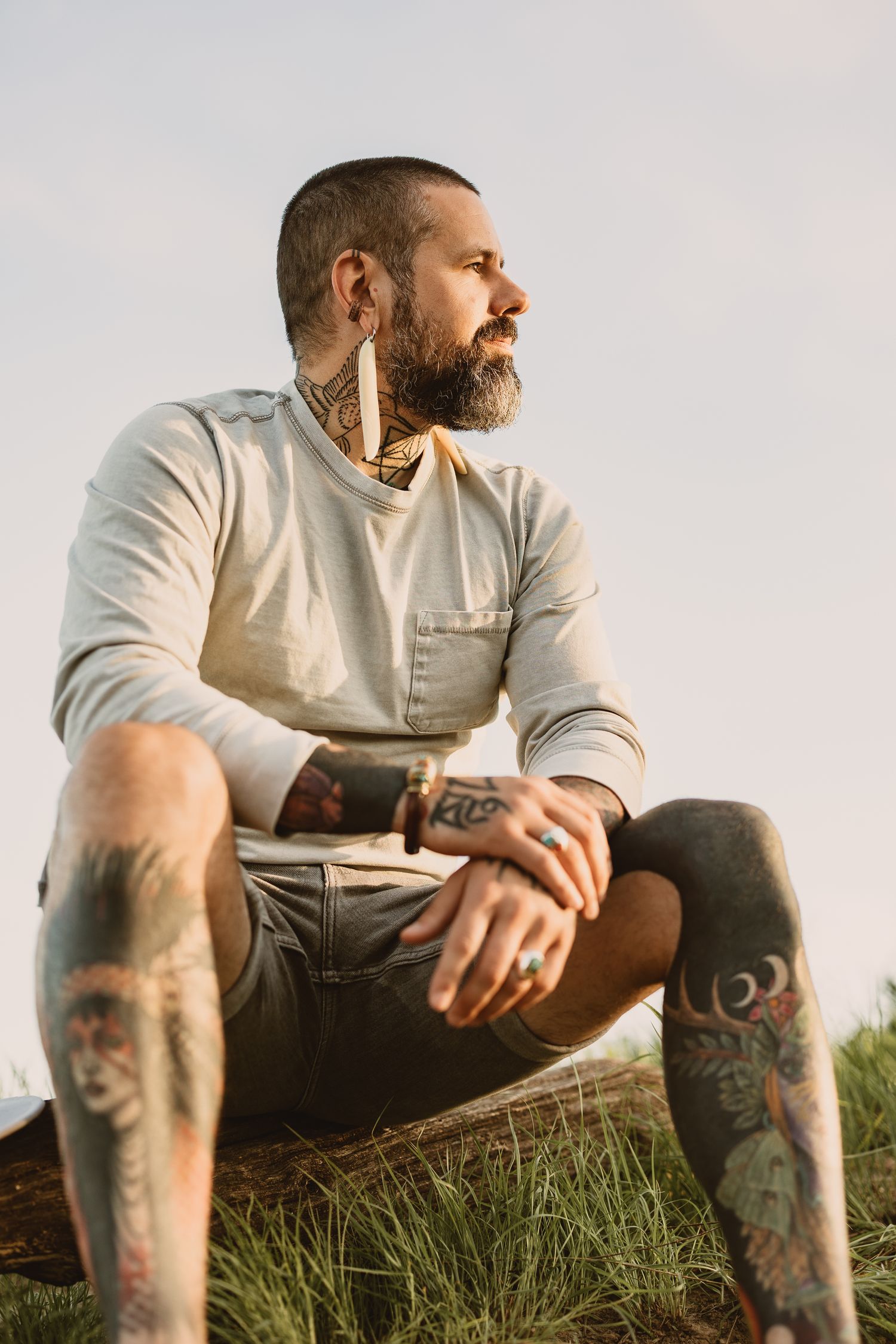 Tattooed bearded man sitting outdoors on grass, wearing casual shirt and shorts, looking away thoughtfully.