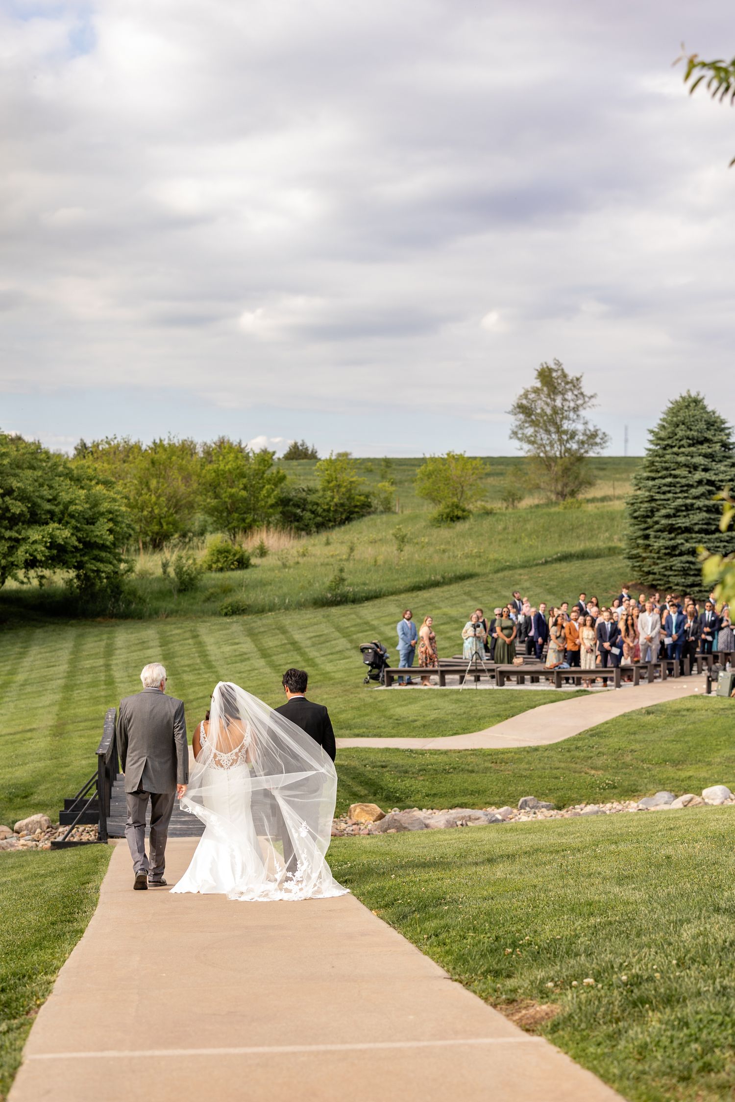 Bride in flowing veil walks down outdoor path with escorts toward wedding guests gathered on a scenic green hillside.