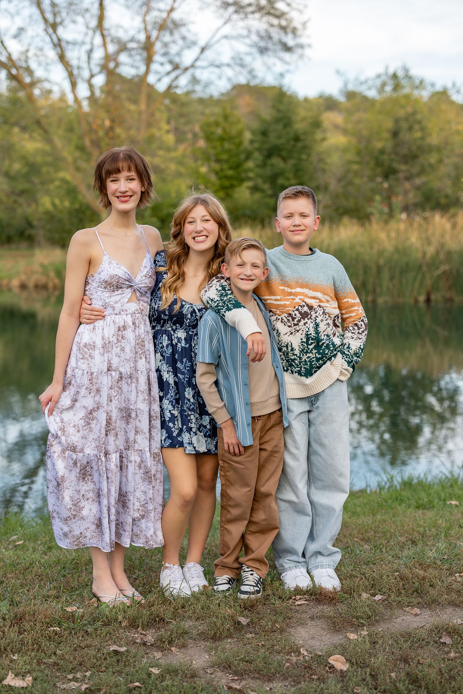 Four smiling siblings pose together near a pond surrounded by autumn trees in a warm outdoor family portrait.