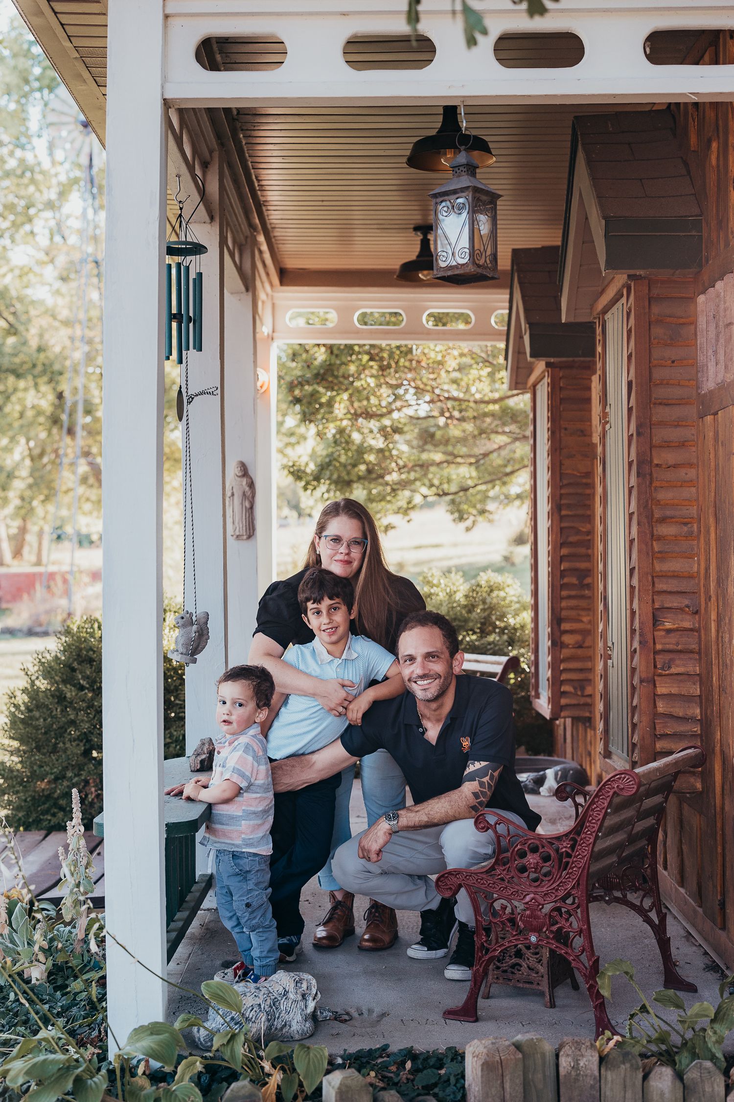 Family of four posing together on a charming rustic porch with red iron chair and lantern light.