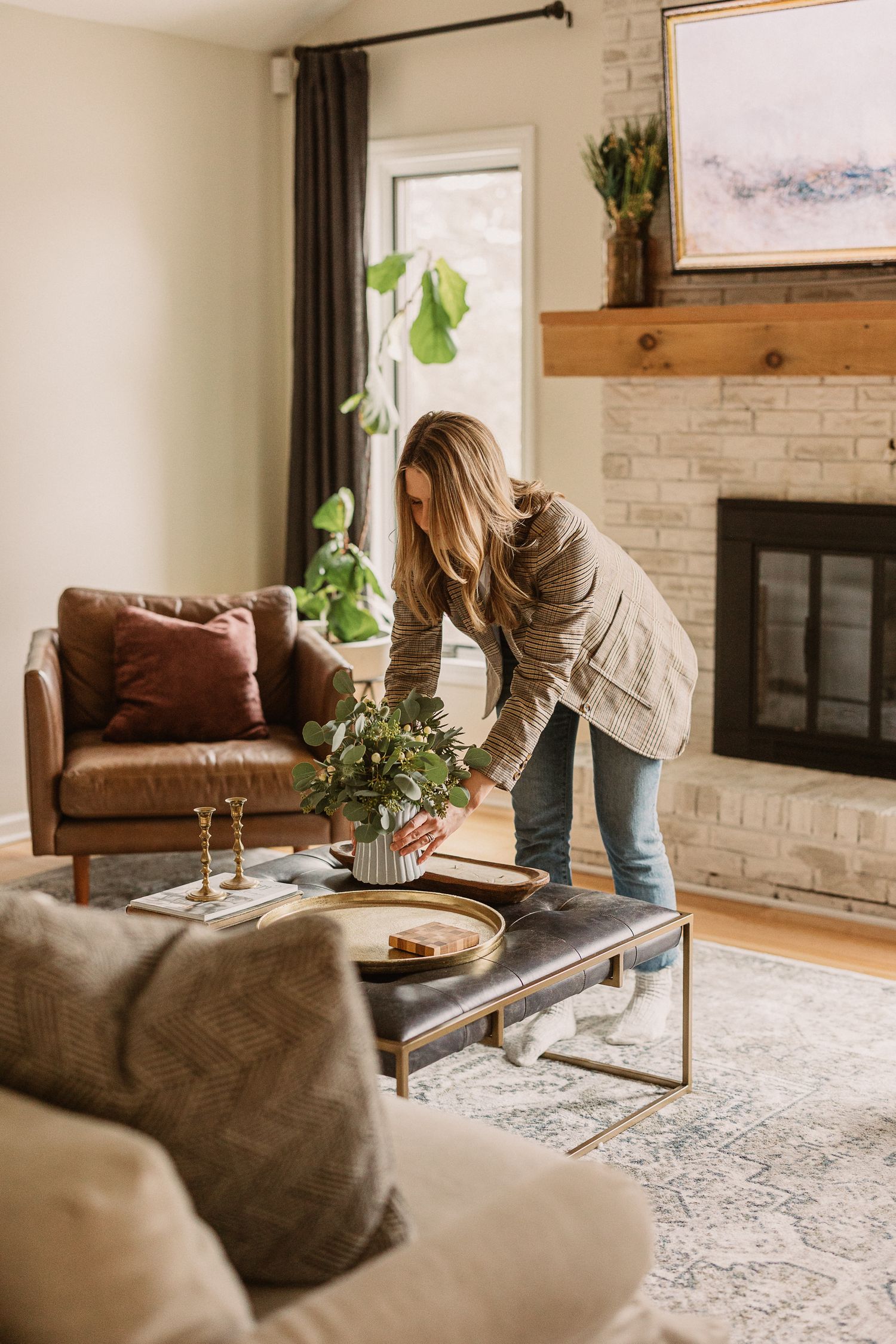 Woman arranging green plant centerpiece on coffee table in cozy living room with leather sofa and stone fireplace.