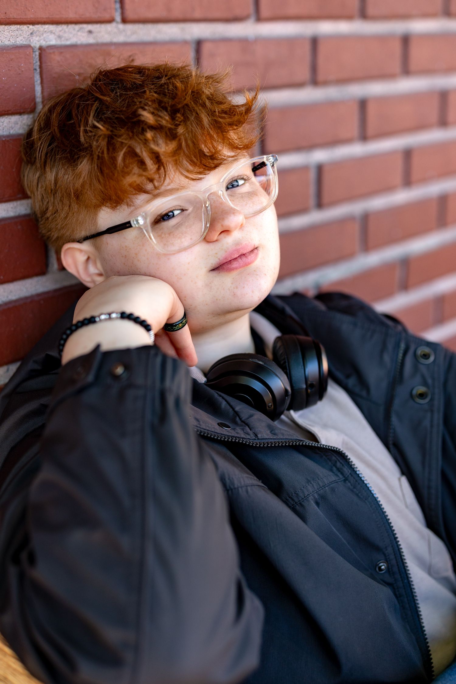 Teenage boy with red curly hair, clear glasses, and black headphones leaning against a brick wall.