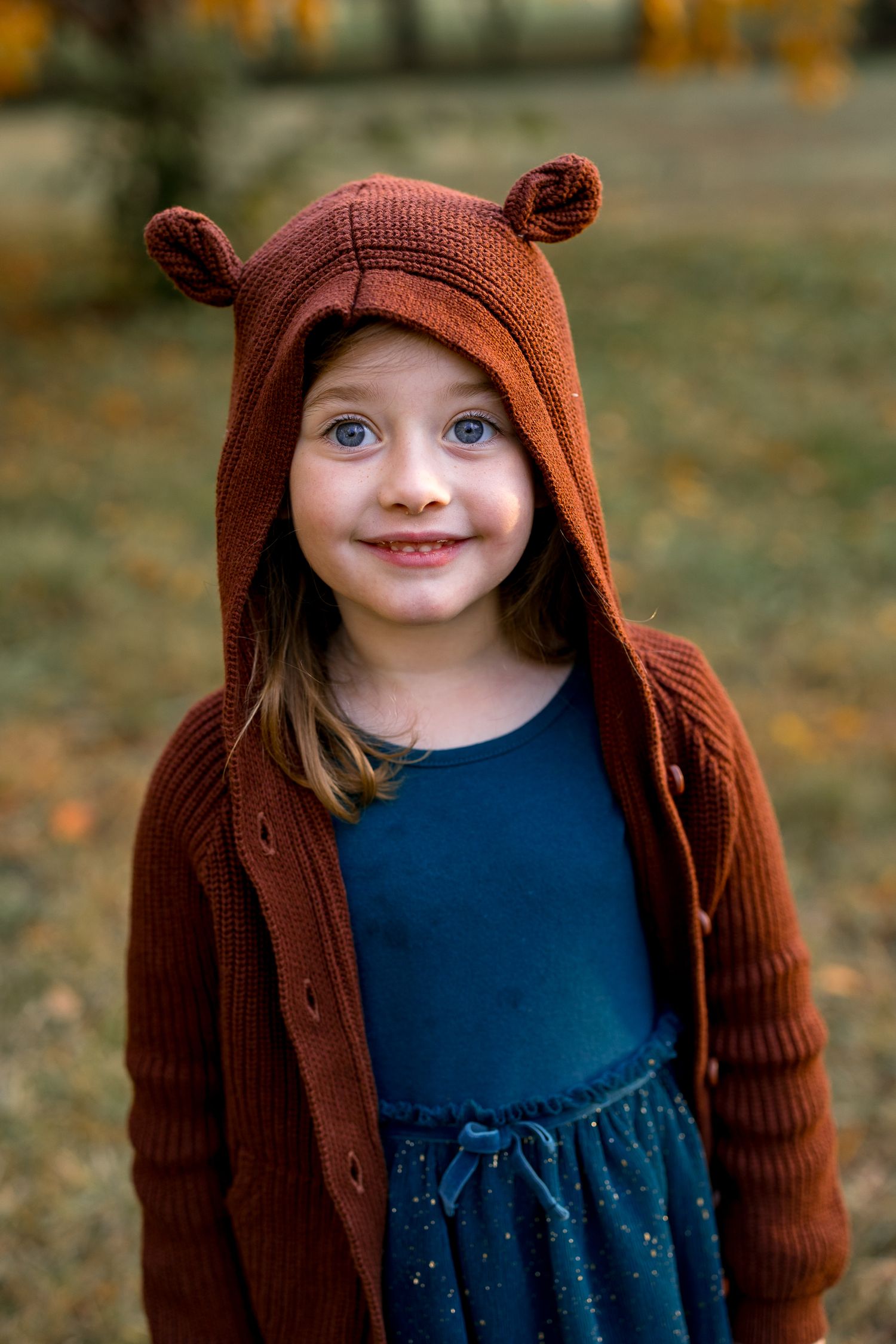 Smiling young girl wearing a brown knit bear ear hoodie cardigan outdoors in an autumn park setting.