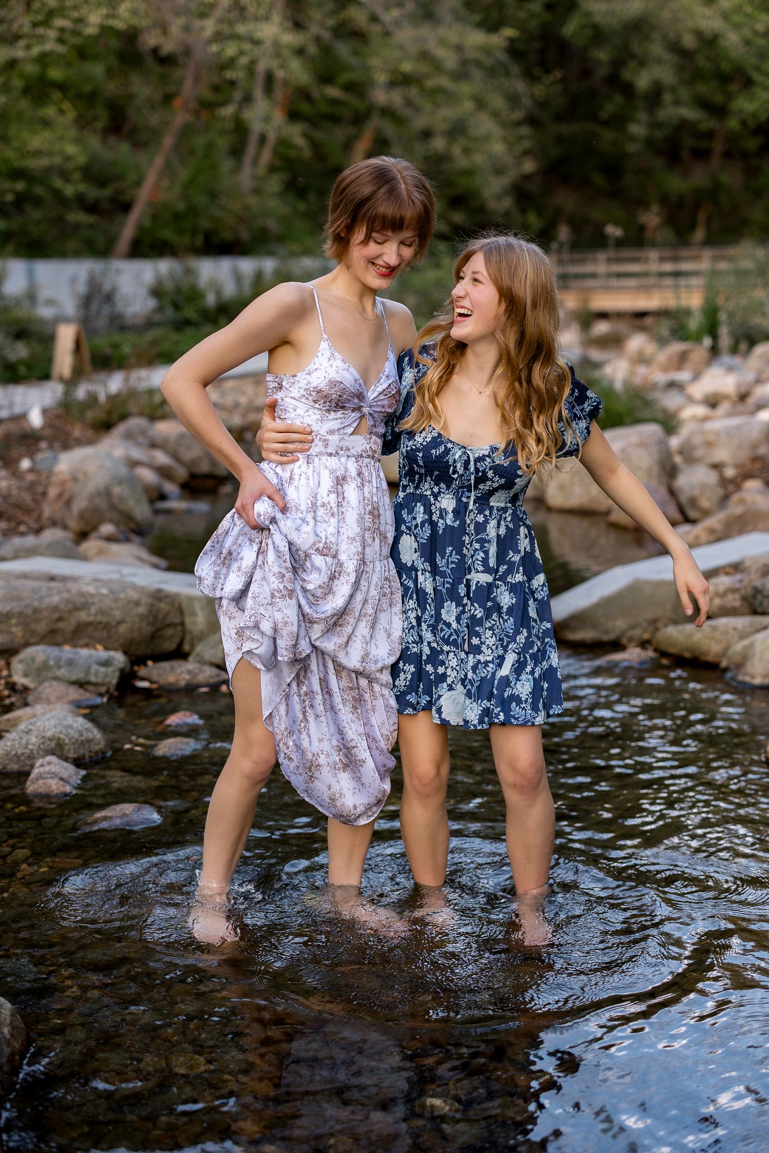 Two young women laugh and wade through a rocky creek together wearing floral dresses outdoors.