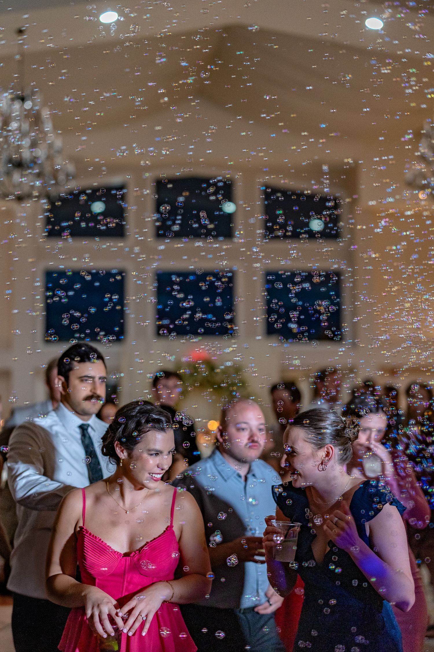 Guests dance joyfully surrounded by floating bubbles at an elegant indoor wedding reception celebration.