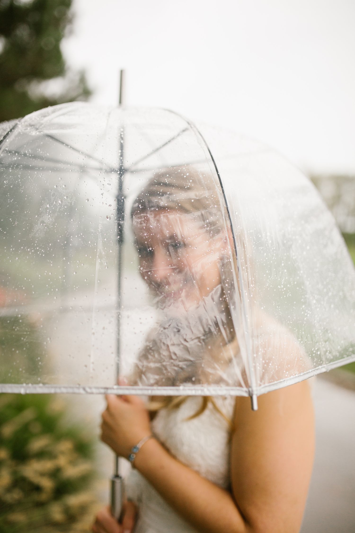 Bride smiling under a clear bubble umbrella on a rainy wedding day, wearing a white dress and bracelet.