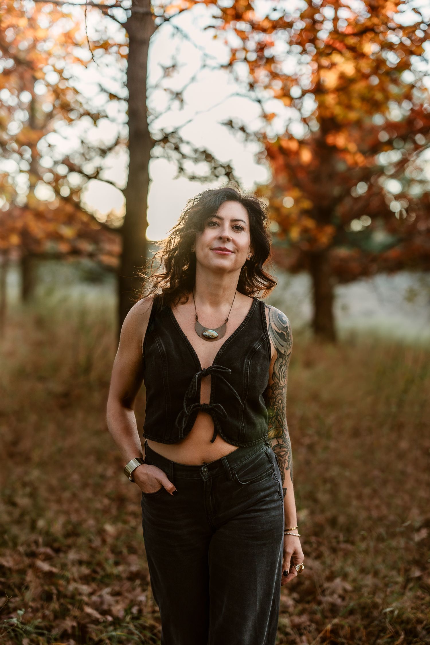Tattooed woman in black crop top and jeans standing in autumn forest at golden hour sunset.