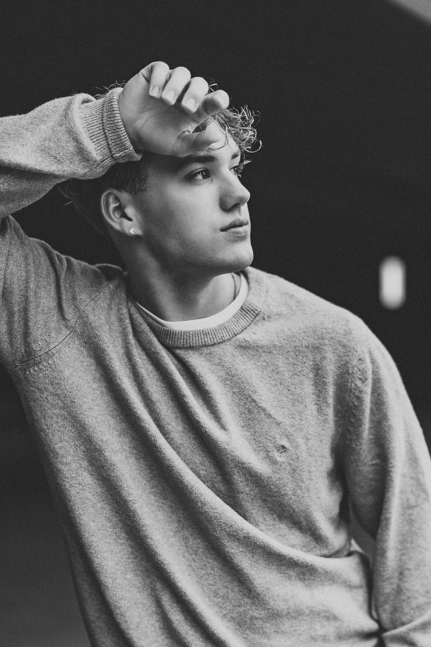 Black and white portrait of a young man in a sweatshirt, hand on forehead, gazing thoughtfully to the side.