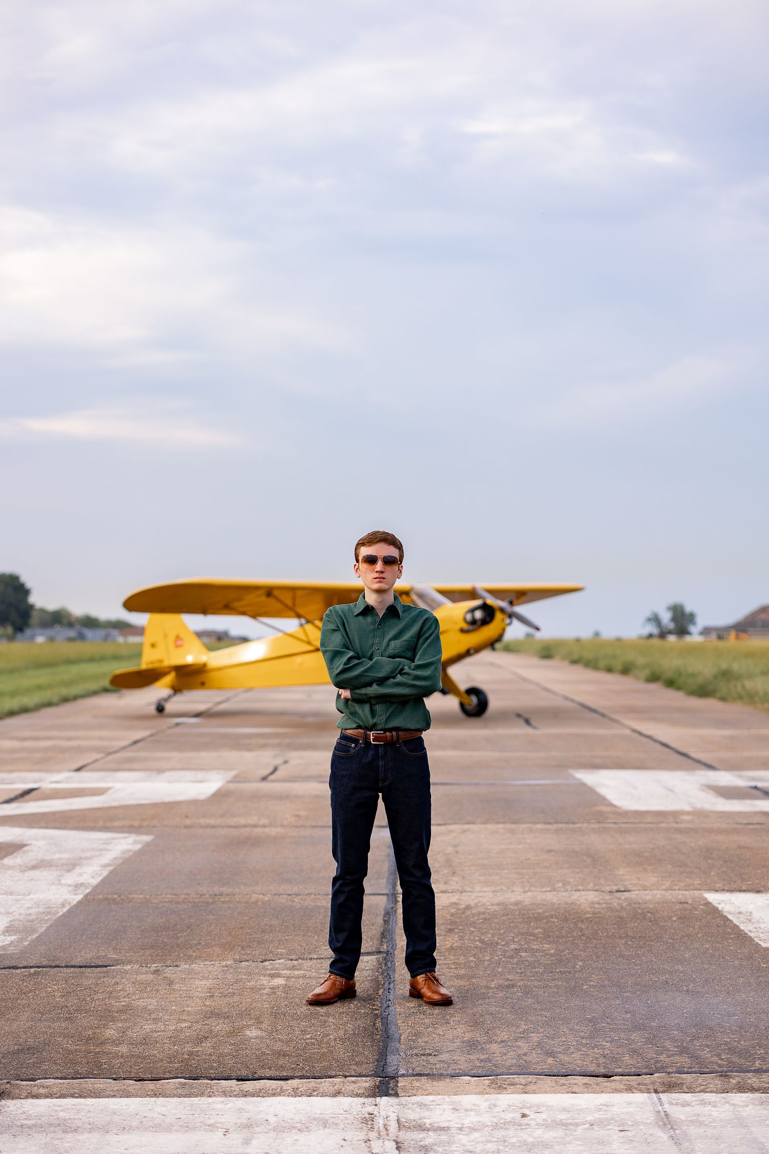 Young man in green shirt and sunglasses stands confidently on runway with yellow small aircraft behind him.