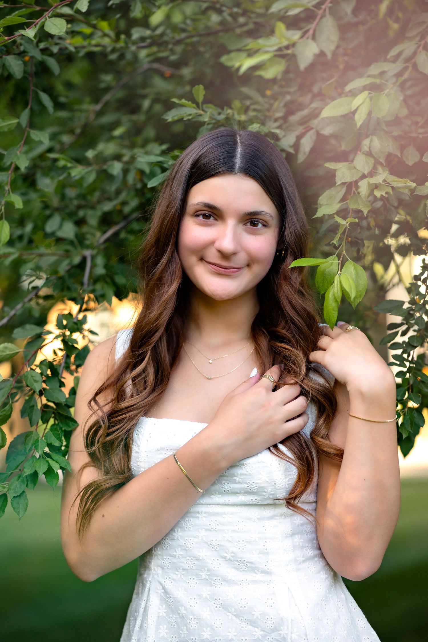 Young woman in white dress smiling outdoors among green leaves, with long wavy brown hair and gold jewelry.