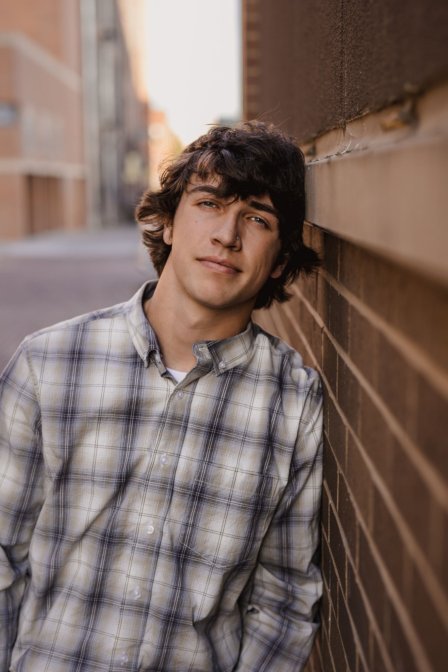 Young man with curly dark hair leans against a brick wall outdoors, wearing a plaid shirt in a candid portrait.