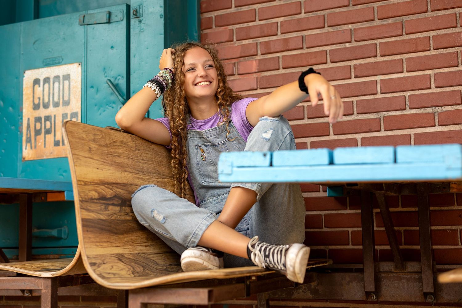 Smiling teenage girl with long braids sits relaxed on a wooden bench near a brick wall and blue table outdoors.
