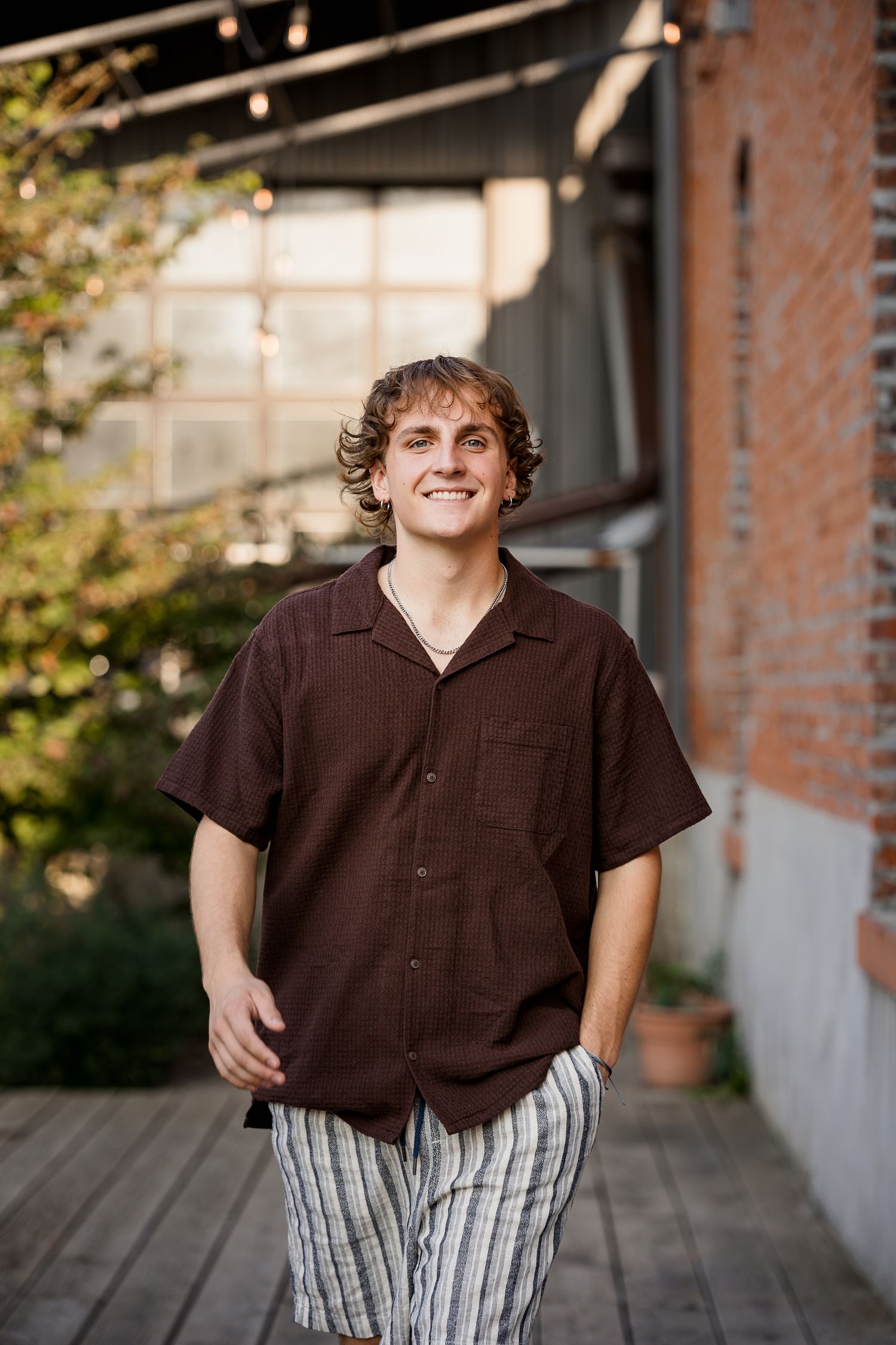 Smiling young man with curly hair wearing a brown shirt and striped shorts standing outdoors near a brick building.
