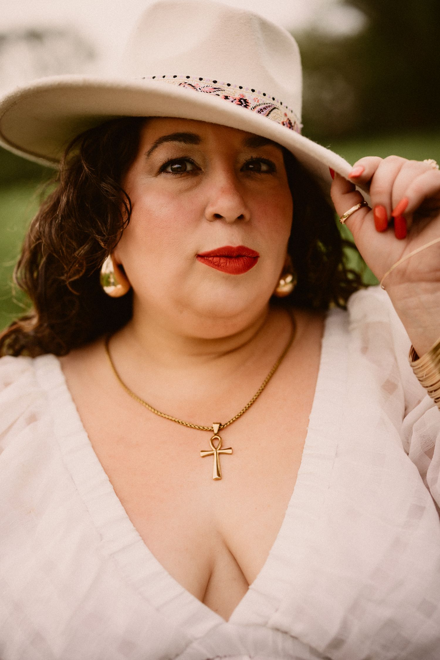 Woman in white hat and dress wearing gold ankh necklace with red lipstick posing outdoors.