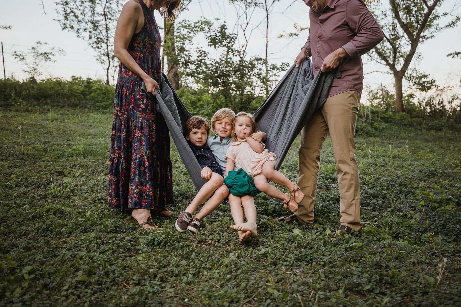 Family of four with two adults holding a gray blanket swing with two young children sitting in it outdoors.