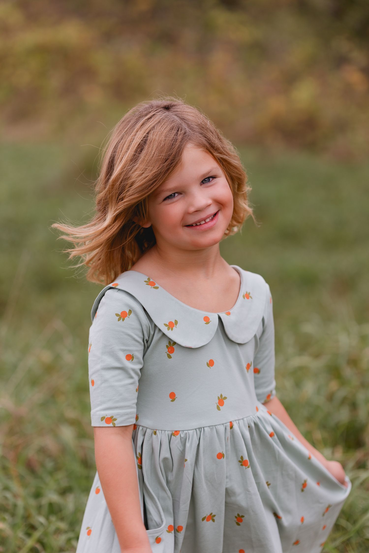 Smiling young girl in a sage green floral dress posing outdoors in a grassy field.