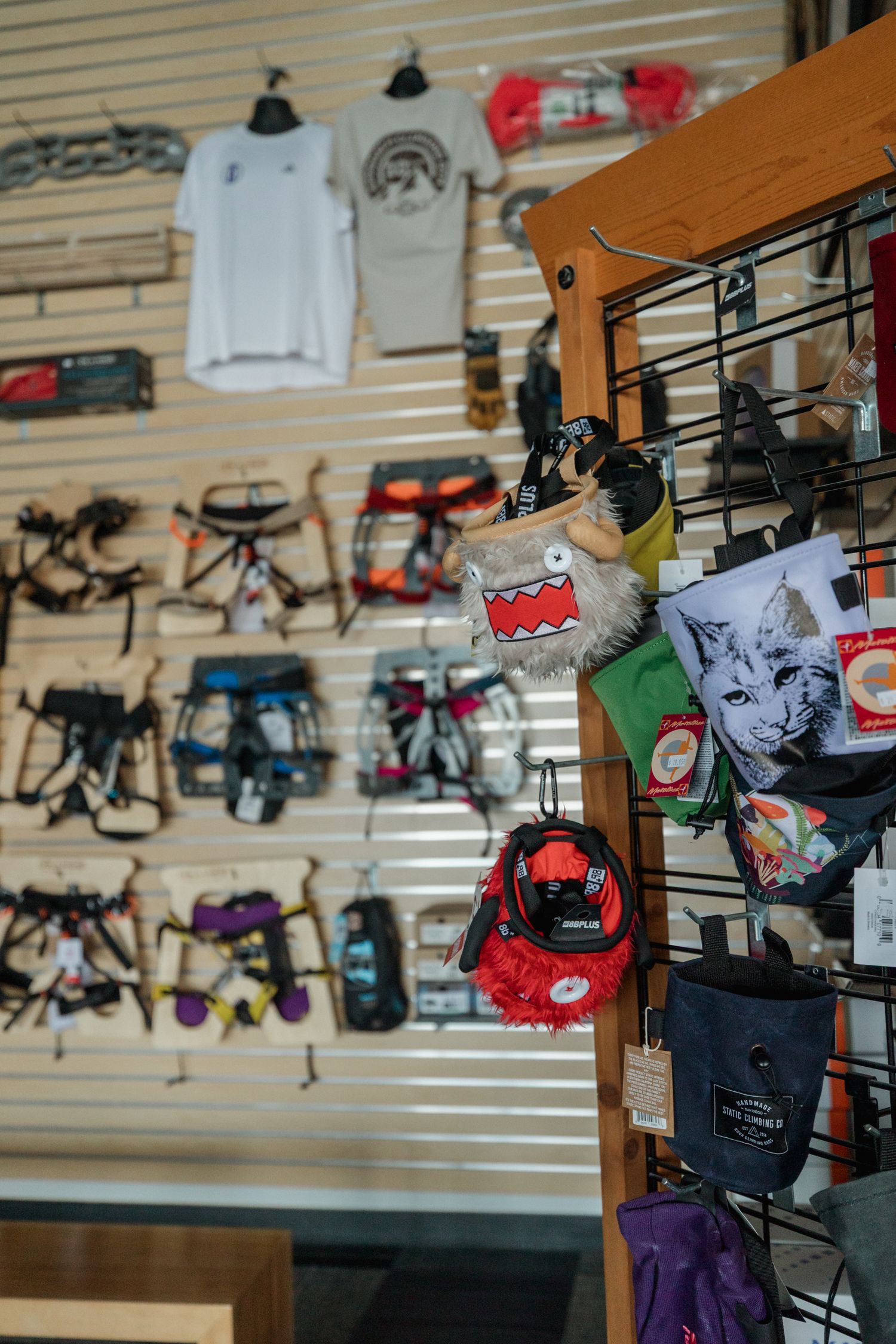 Outdoor gear shop display with climbing harnesses, t-shirts, and novelty bags including a Domo character on a retail rack.