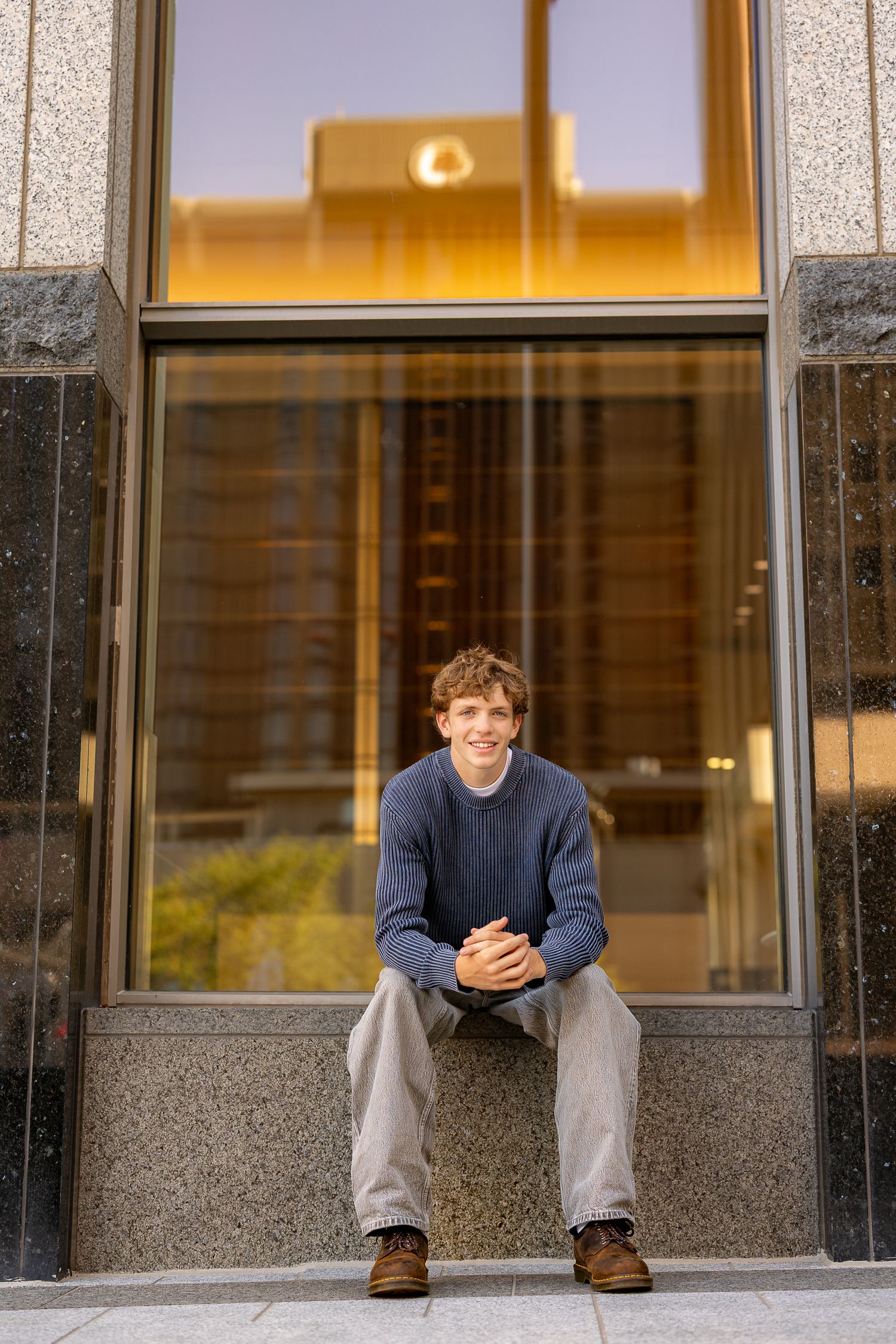 Young man in blue sweater and khakis sitting on granite ledge outside modern glass building, smiling confidently.