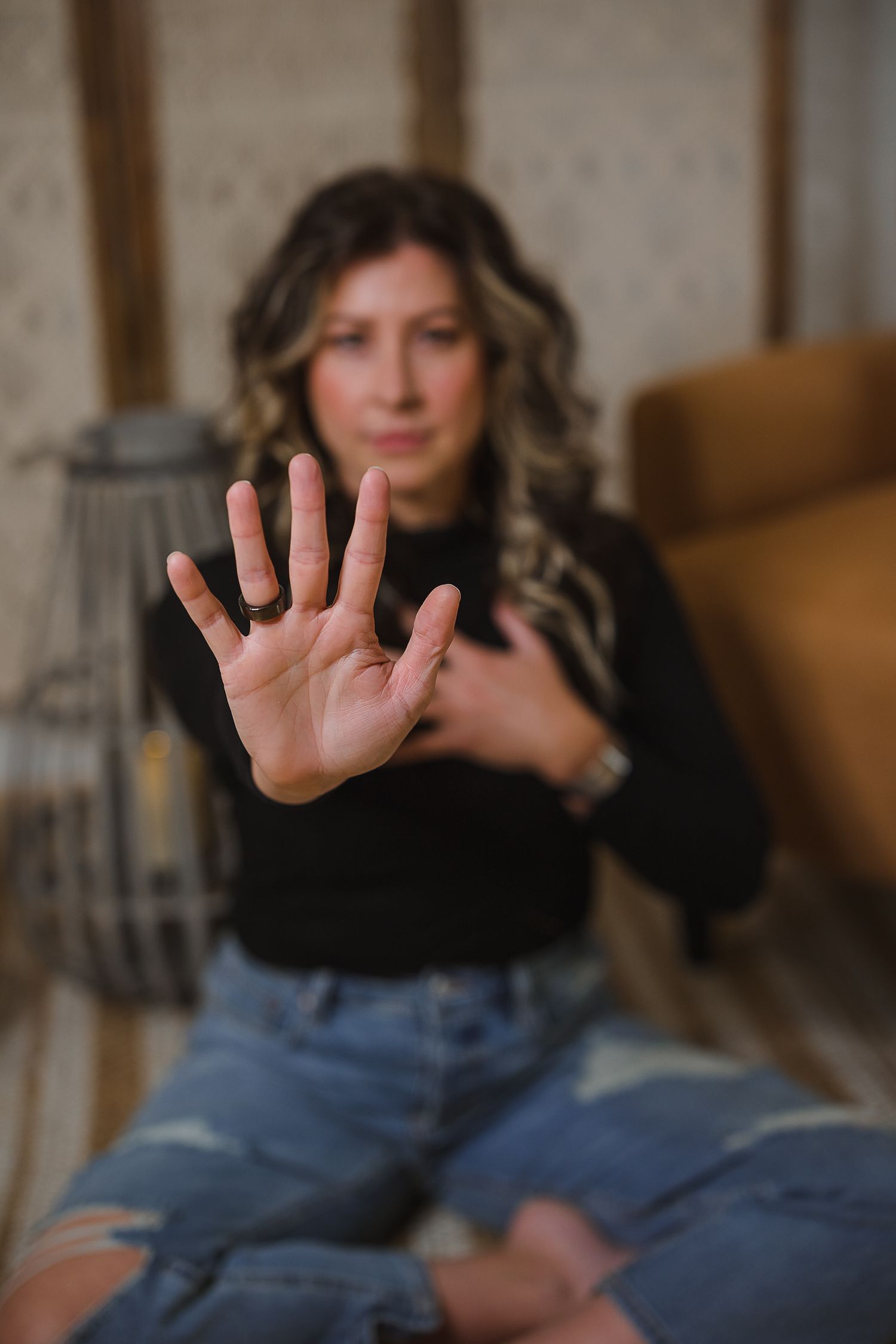 Woman in black top and ripped jeans holding hand up in stop gesture while sitting on floor indoors.
