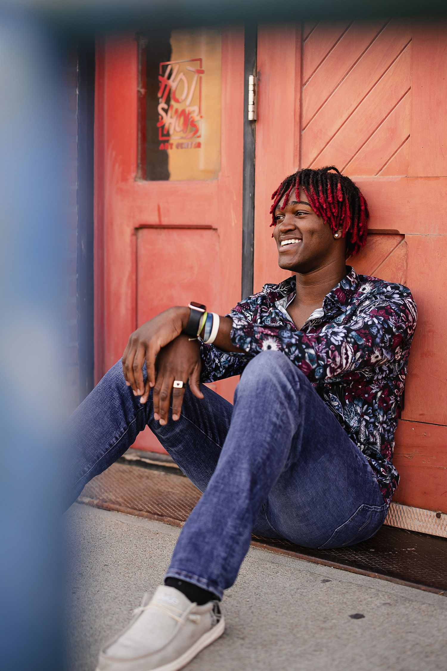 Smiling young man with red dreadlocks sits against a red door wearing a floral shirt and jeans.