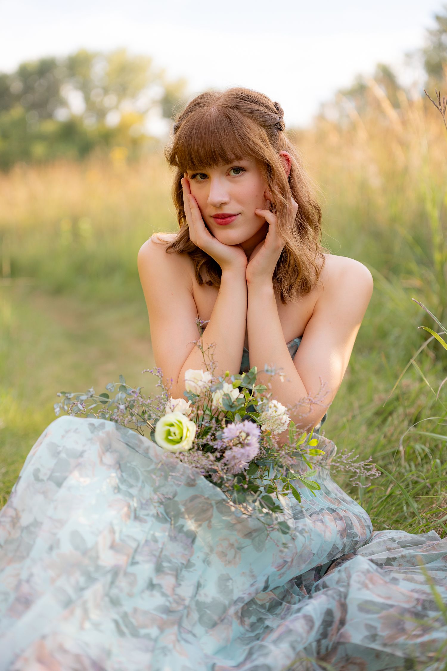 Young woman in floral dress sitting in meadow, holding wildflower bouquet, hands framing face, golden hour light.