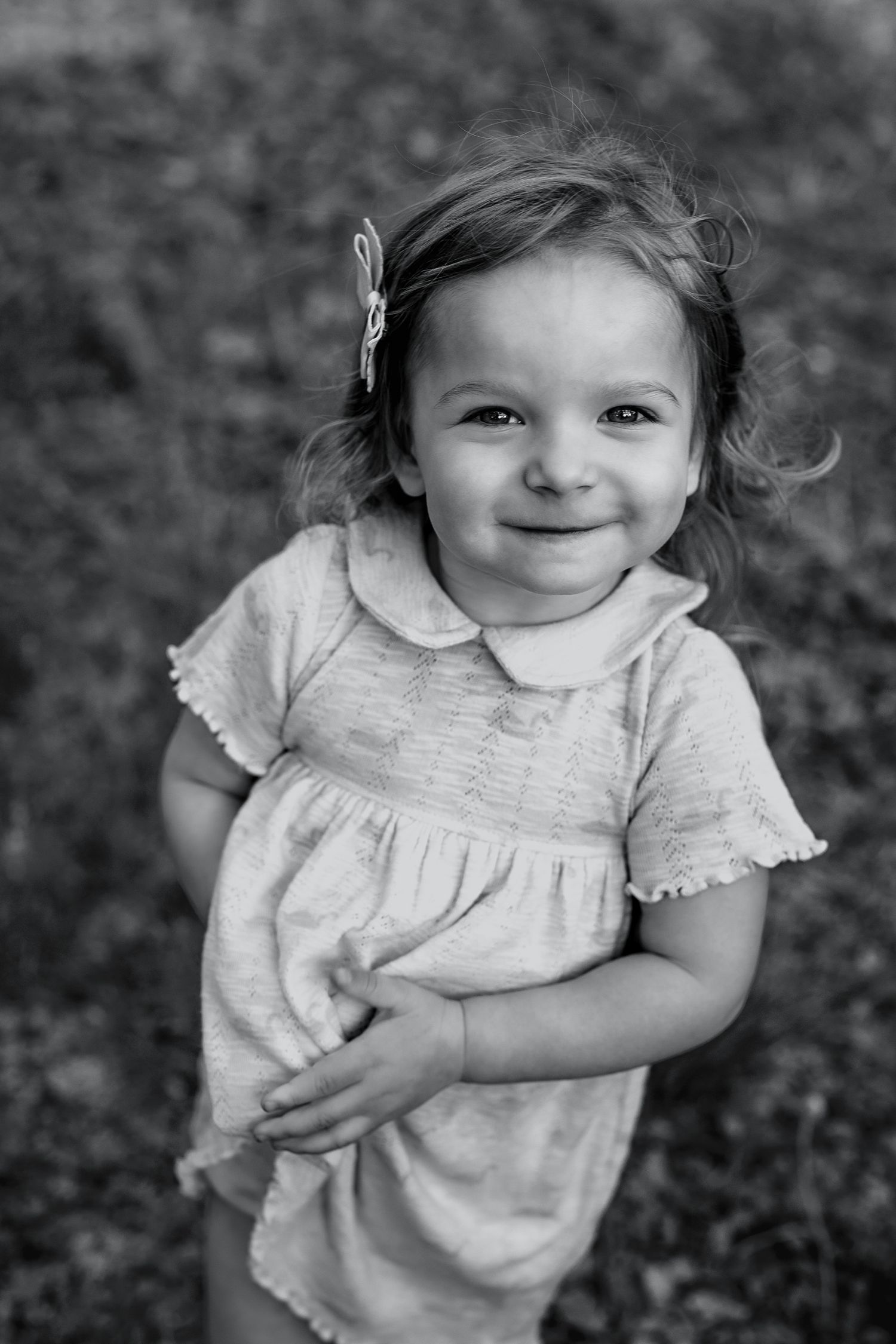Black and white portrait of a smiling toddler girl in a dress with a hair clip, standing outdoors.