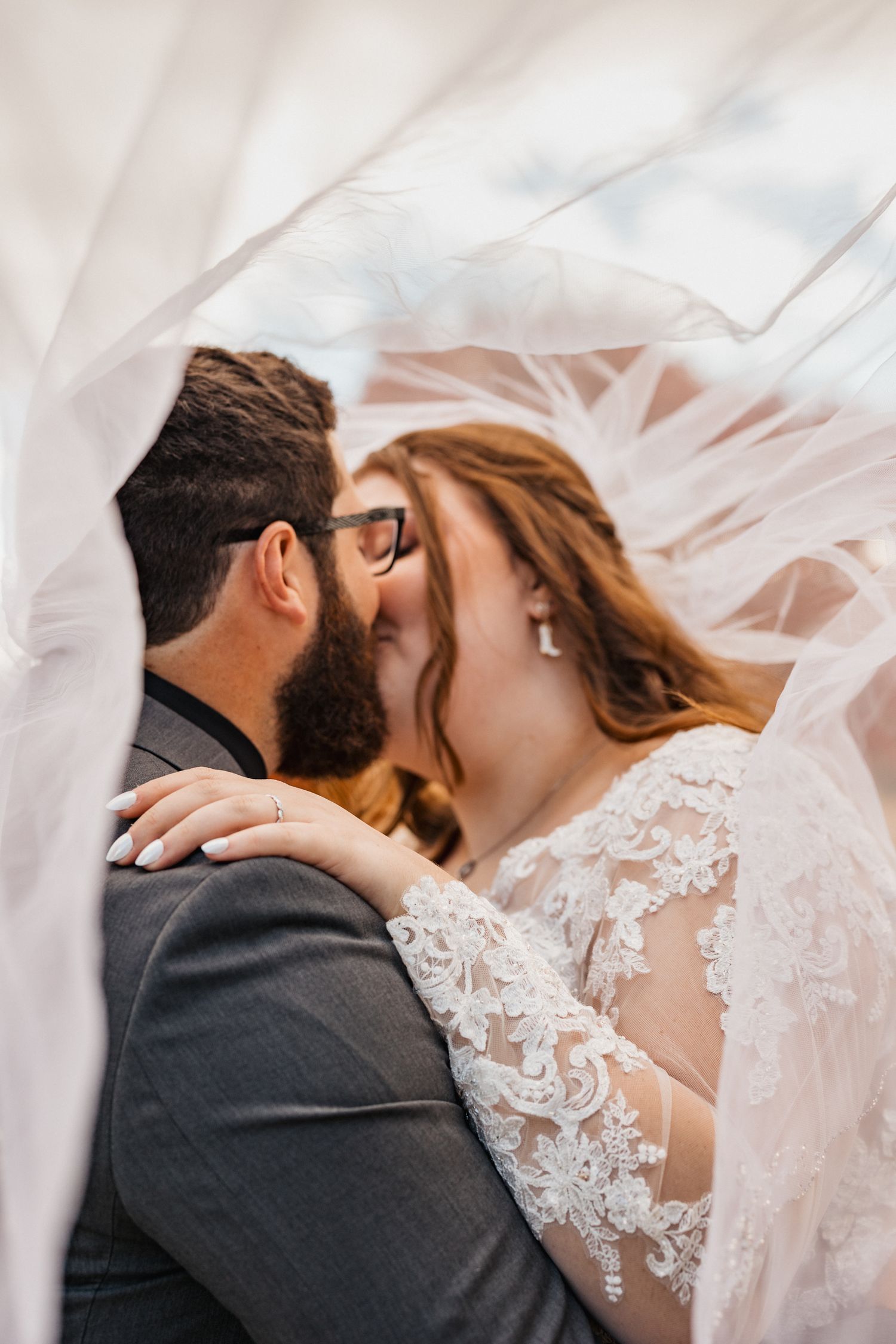 Bride and groom sharing a romantic kiss under a flowing wedding veil, bride wearing lace dress and glasses.