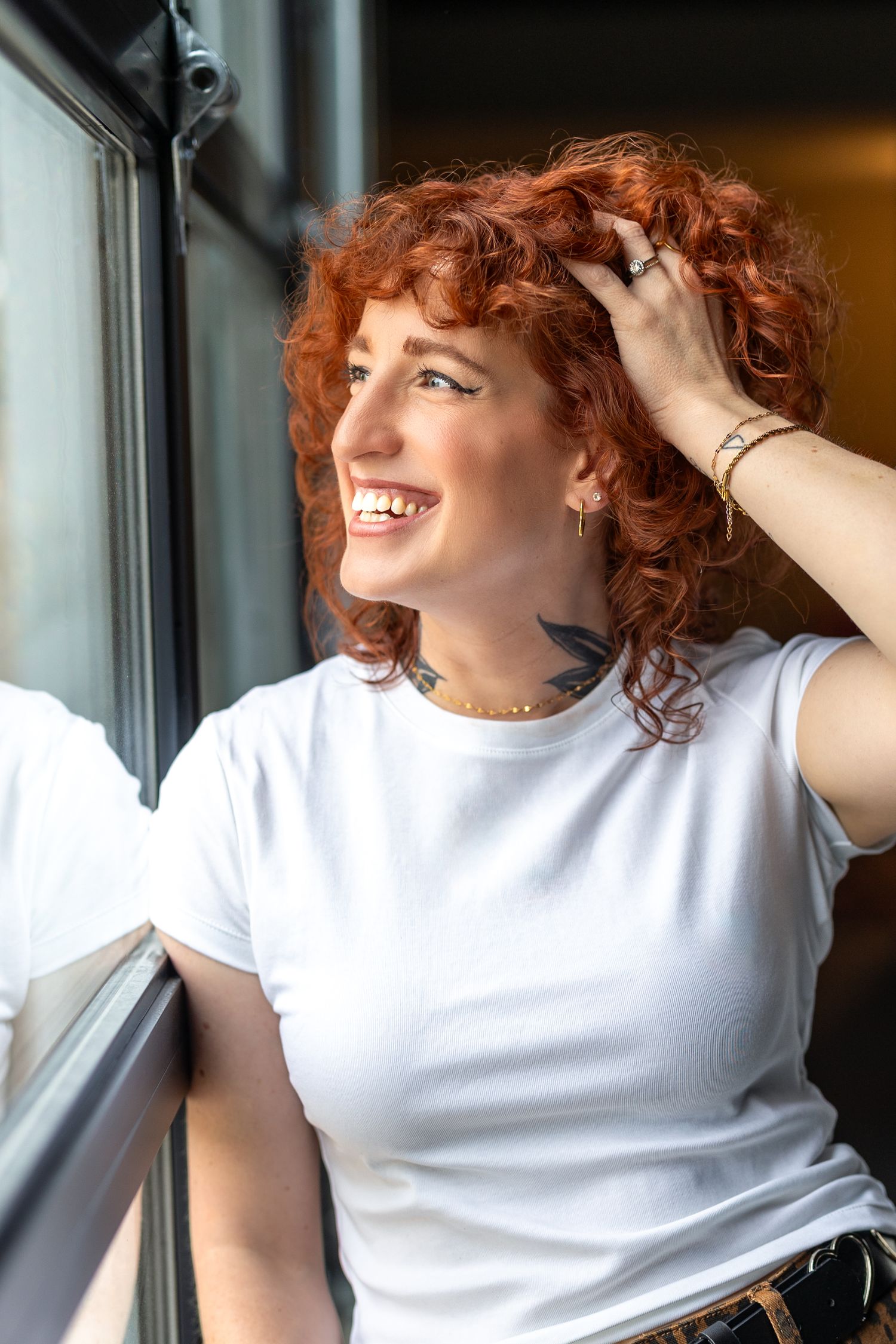 Smiling redhead woman with curly hair, neck tattoo, and white t-shirt, posing by a window in natural light.