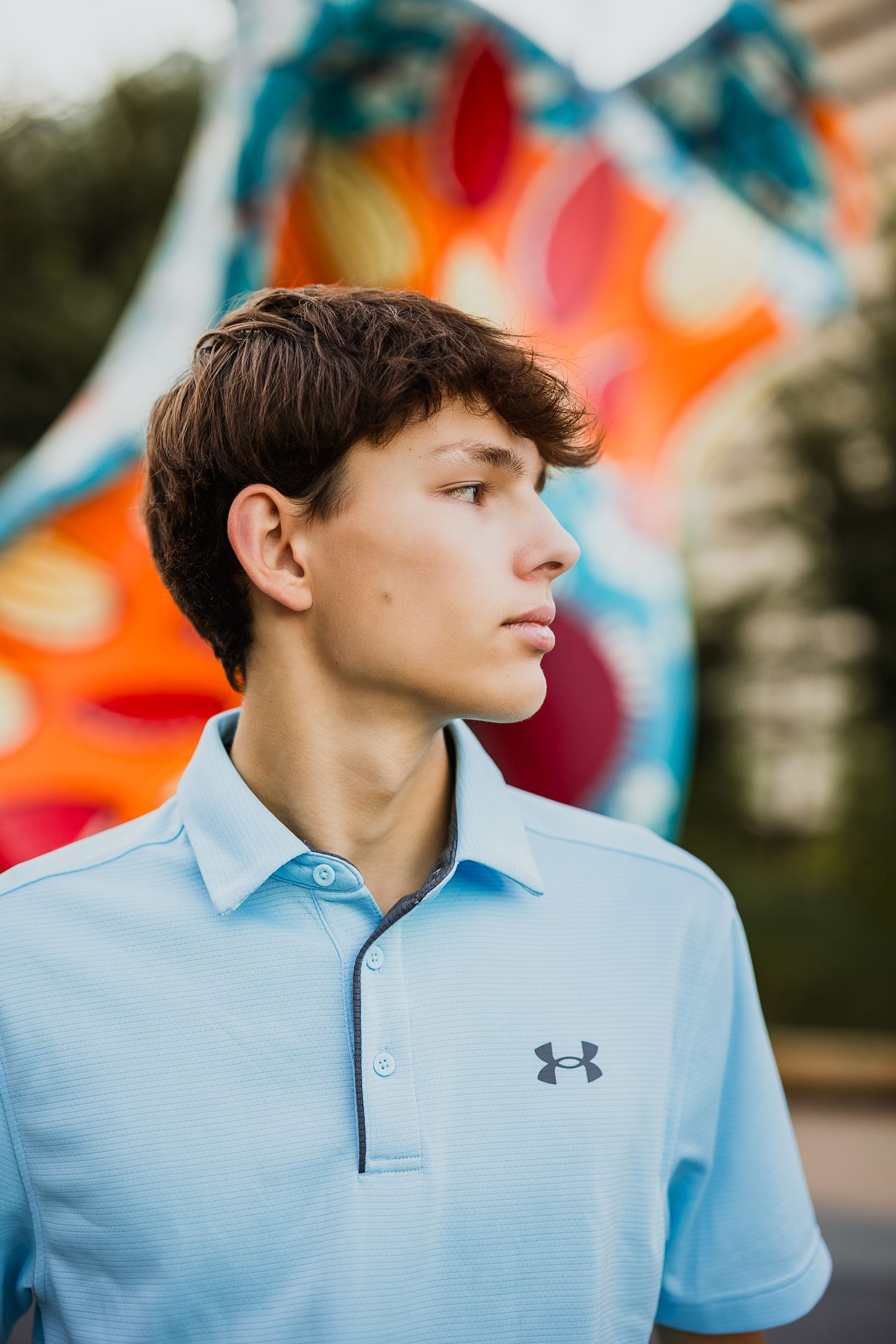 Teen boy in light blue Under Armour polo shirt standing outdoors with colorful mural in background.